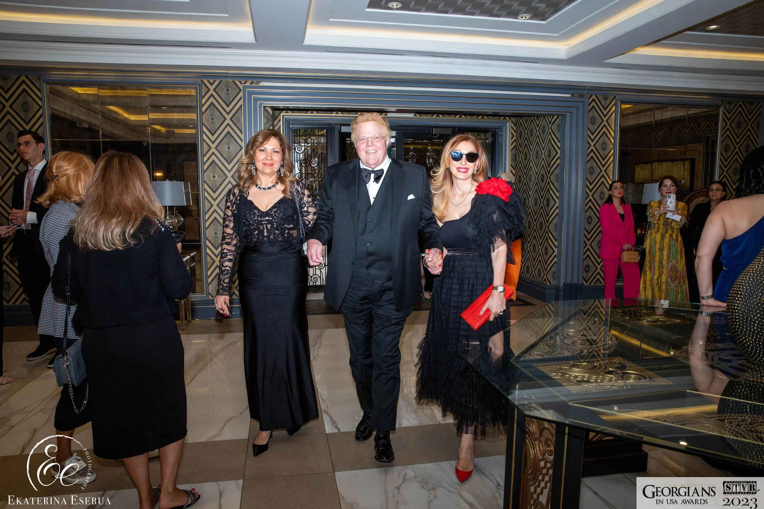 People dressed in formal attire at a luxury event, with three individuals holding hands and smiling at the center. The background features a patterned wall and mirrors, with other guests visible in the background.