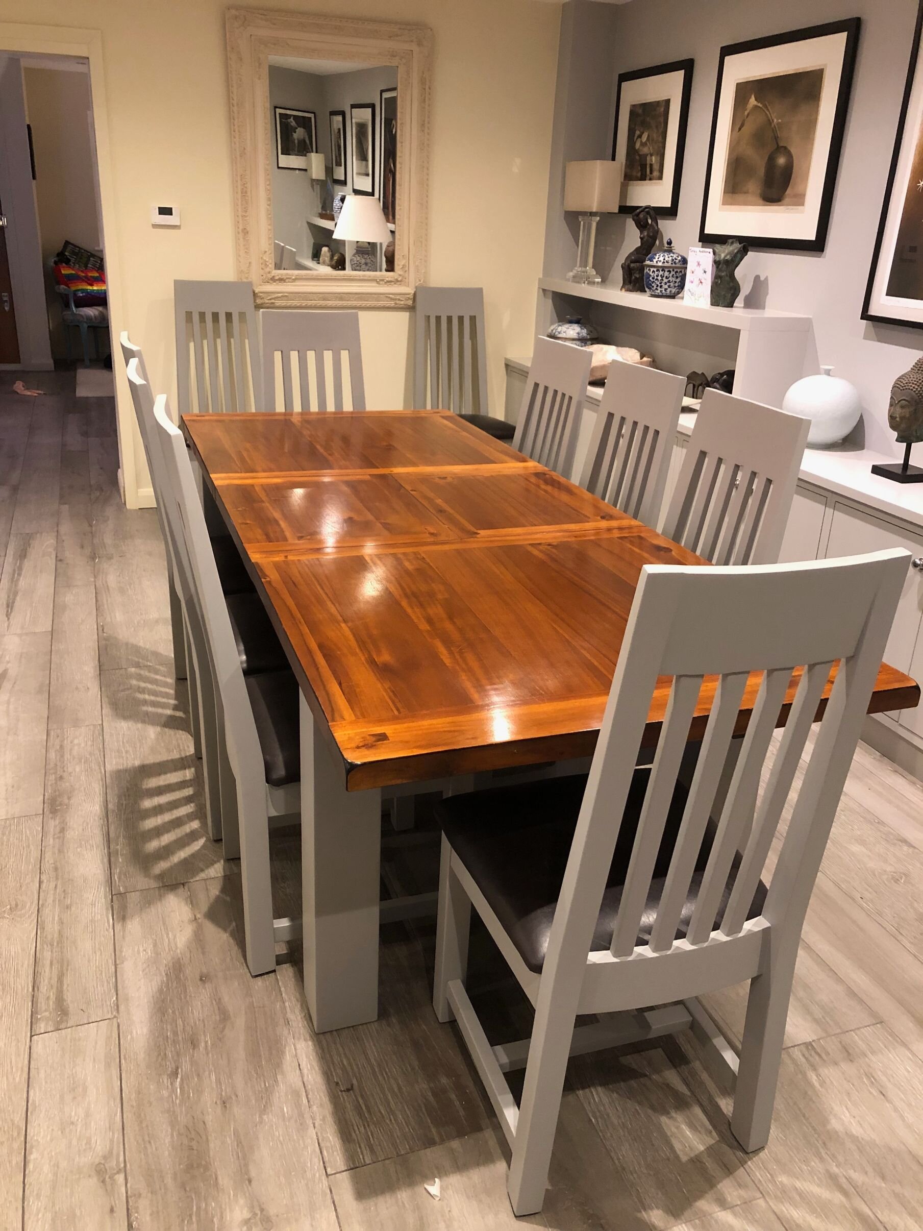 Dining room with a wooden table and gray chairs, framed artwork on the wall, decorative items on a shelf, and a large mirror reflecting the room.