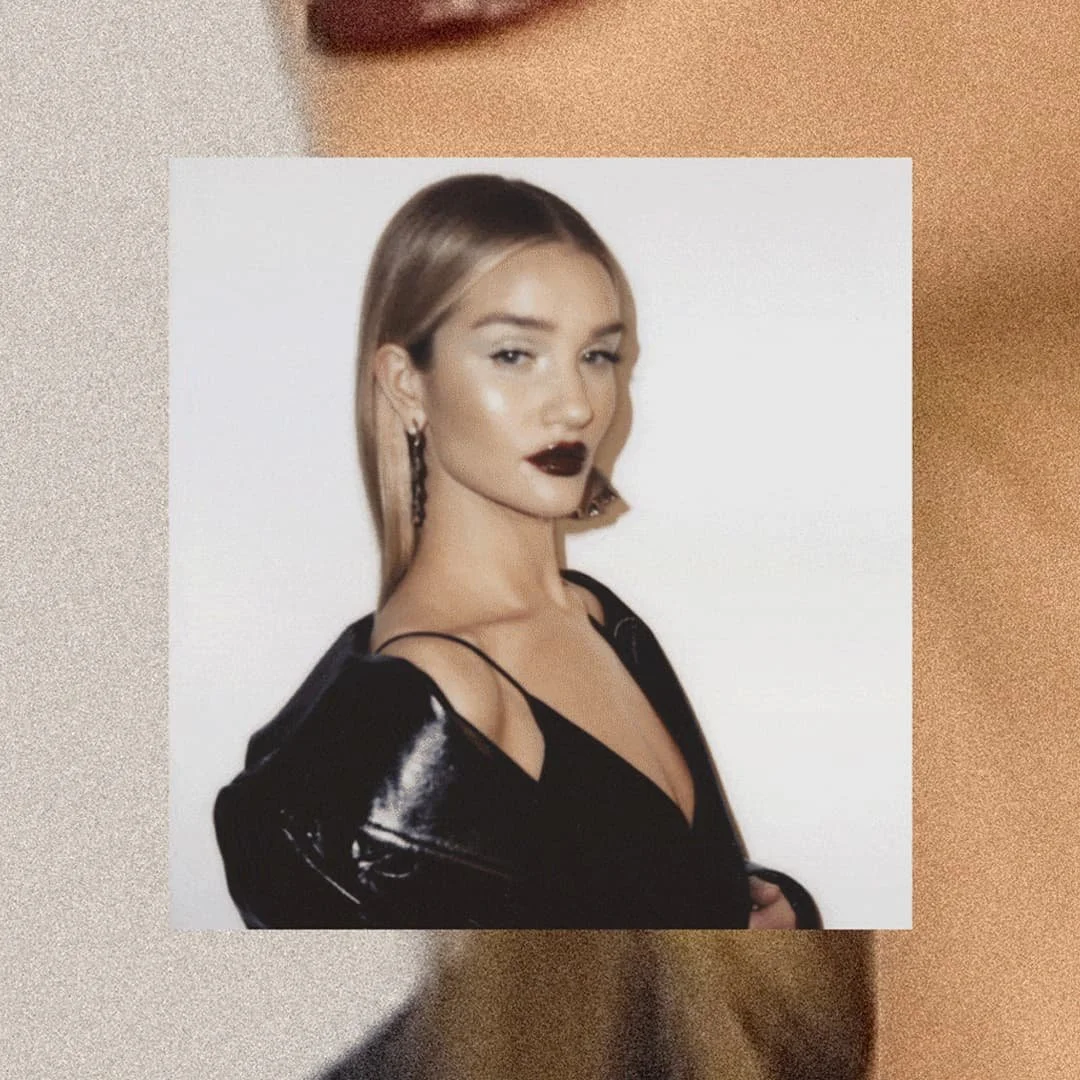 A woman with blonde hair and dark lipstick posing against a white background, wearing a black outfit with thin straps and earrings.