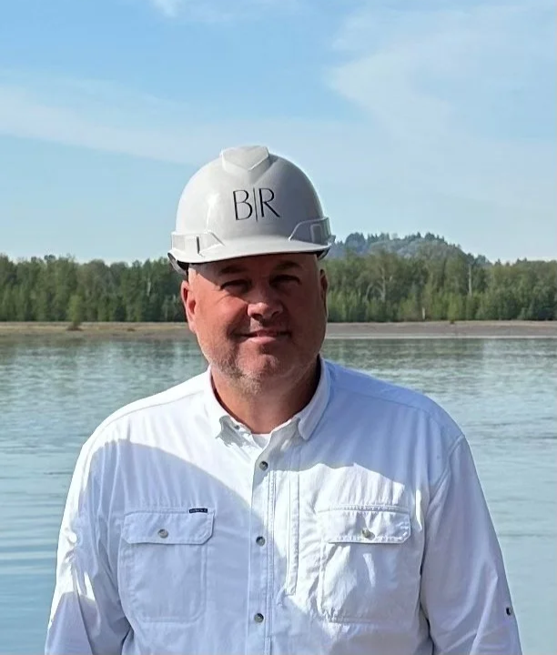 Man wearing a construction helmet with 'BIR' written on it, standing outdoors near a body of water with trees in the background.
