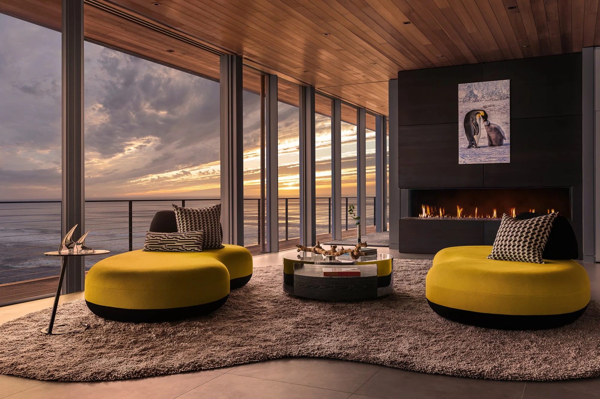 Modern living room with large floor-to-ceiling windows overlooking the ocean during sunset. It features two yellow oval chairs with patterned cushions, a round glass table with decorative driftwood, a black fireplace with a photo of penguins above it, and a plush gray rug.