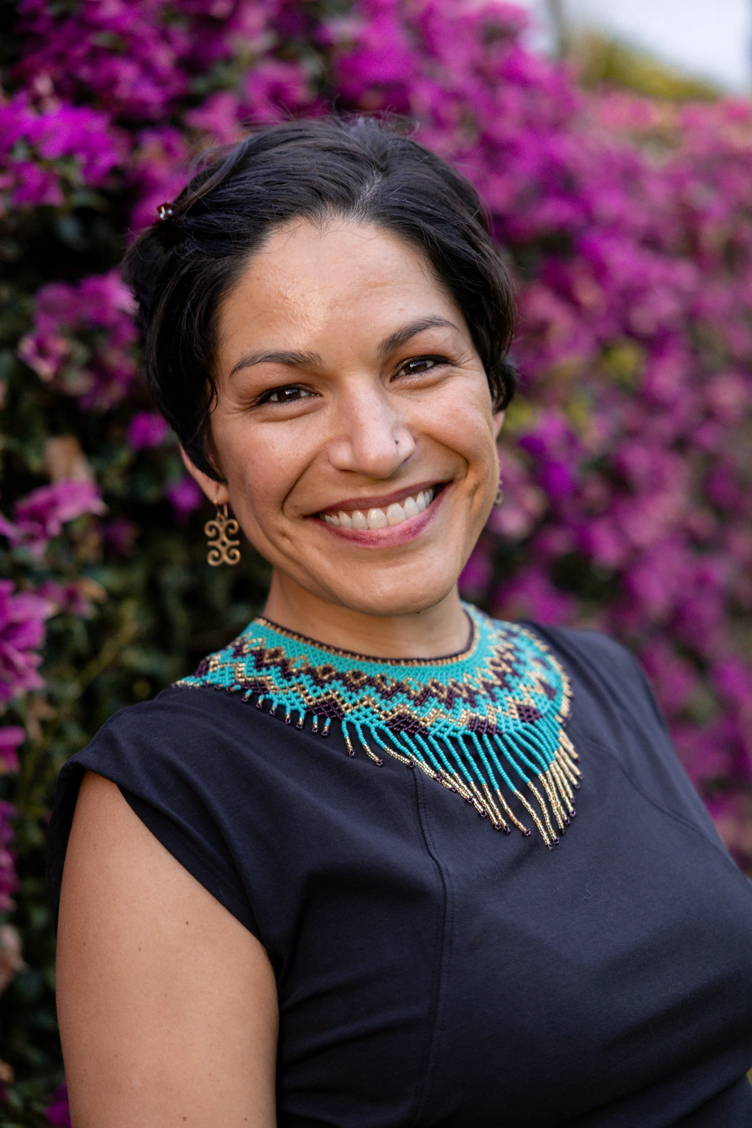 A woman smiling outdoors with pink flowering bushes in the background, wearing a colorful beaded necklace and earrings.