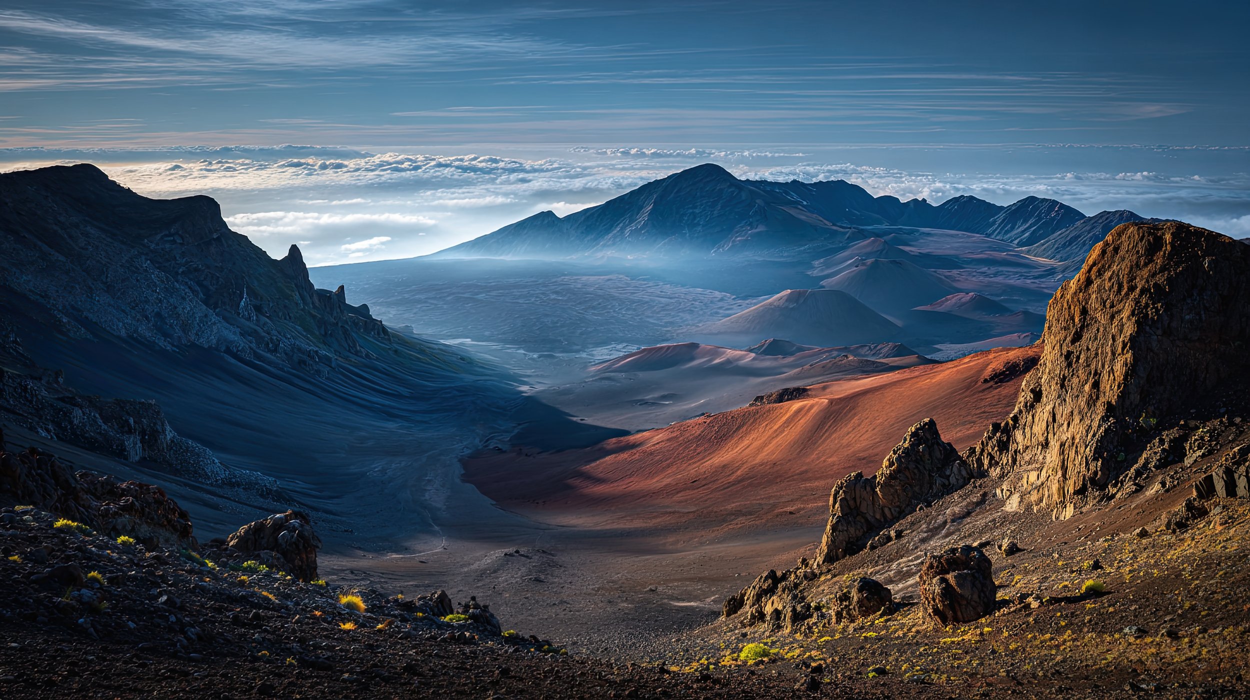 A mountainous landscape with steep rocky slopes and volcanic craters, partly covered in snow and ash, under a partly cloudy sky.