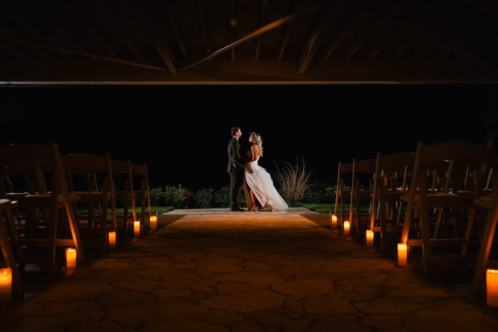 Bride and groom holding hands and looking at each other on a wedding altar at night, with candles lining the aisle and chairs on both sides.