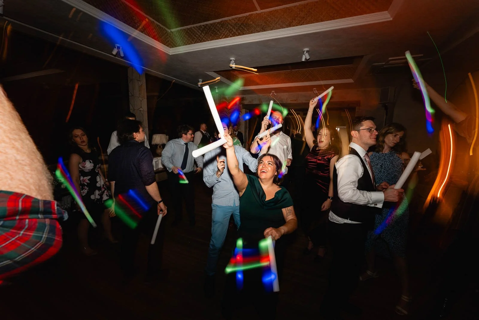 People dancing and celebrating with glow sticks and light trails in a dimly lit party venue.