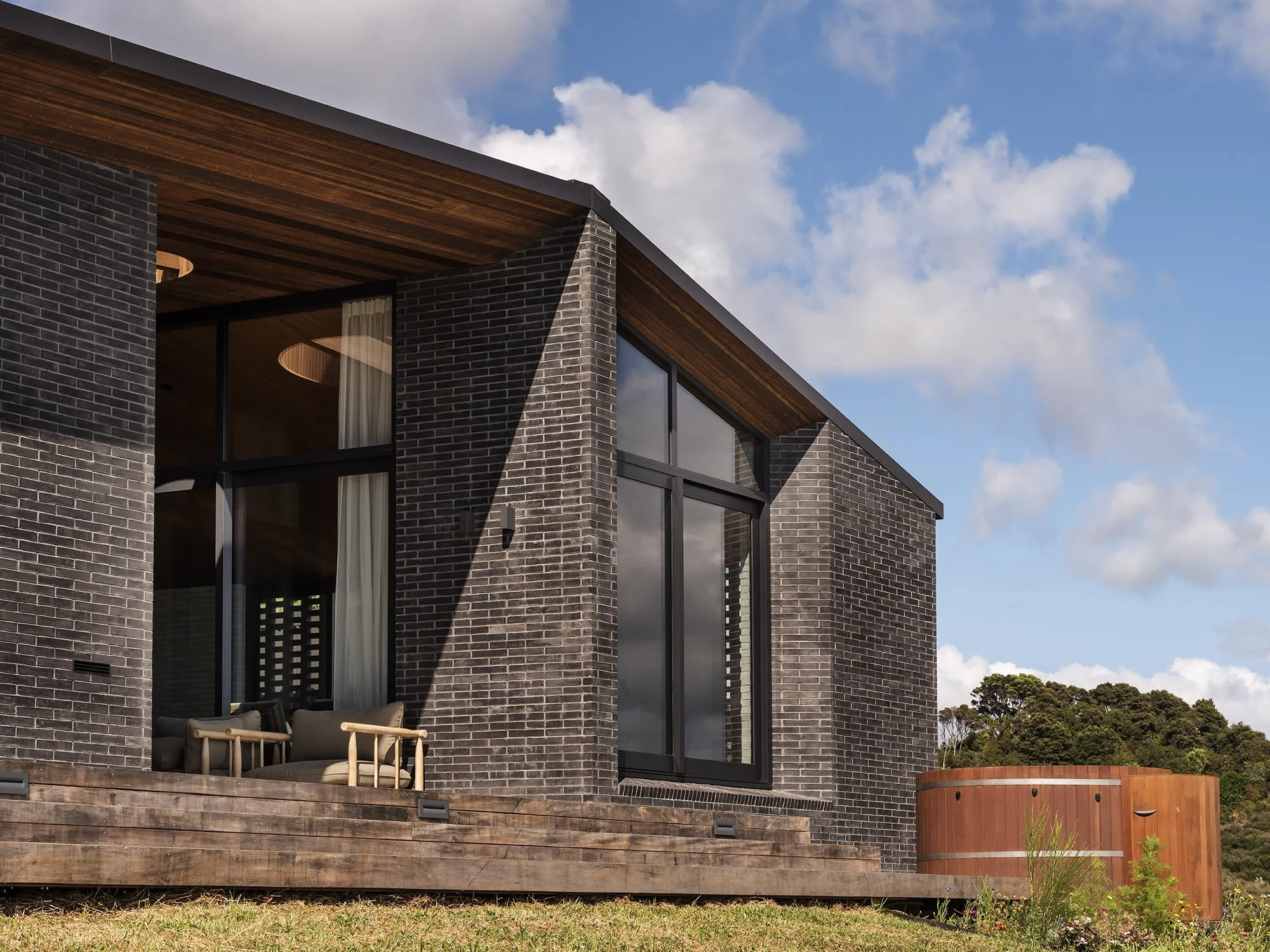 Modern house with brick exterior, large glass windows, wooden deck, outdoor furniture, and a cloudy blue sky.