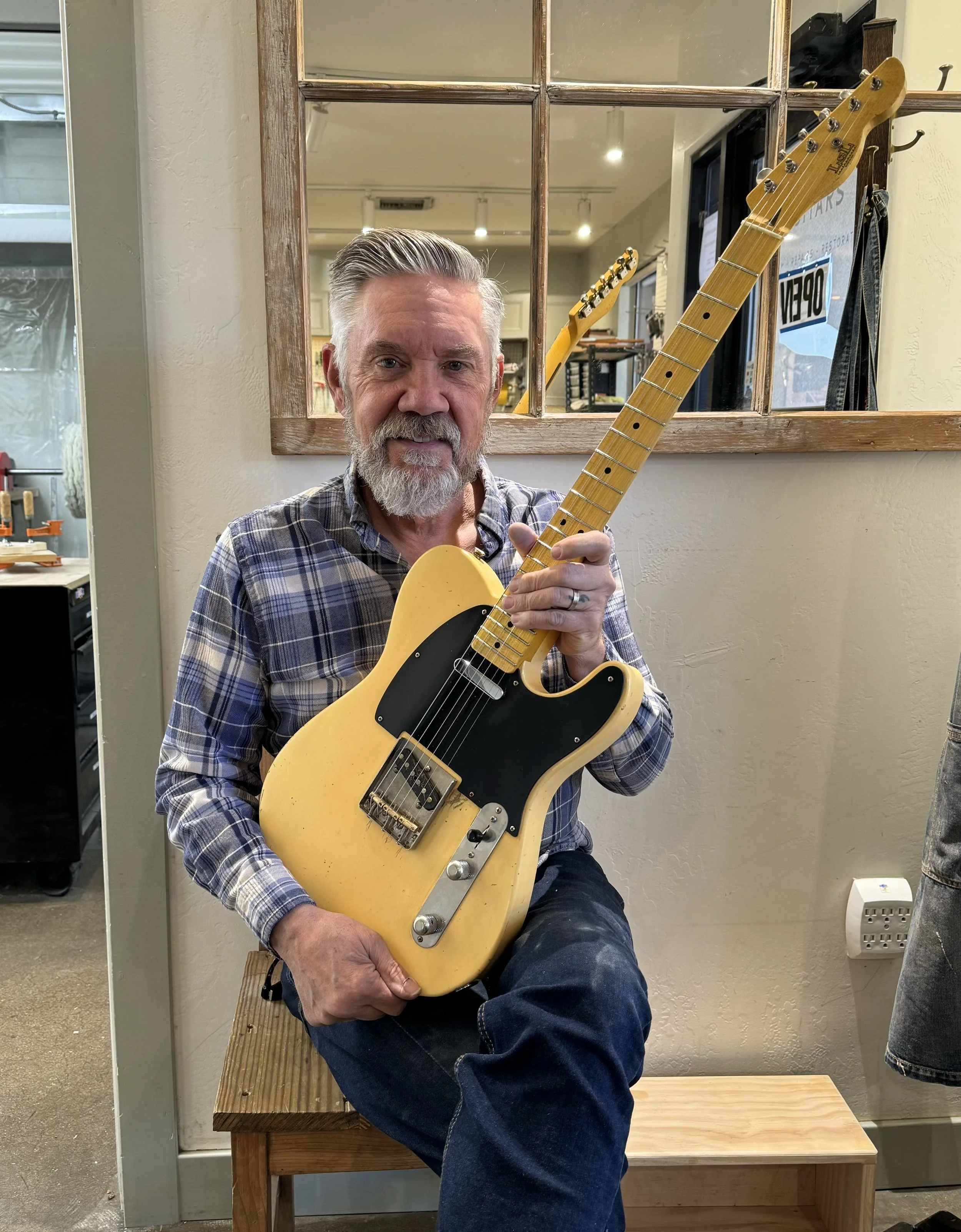 Chris Fleming sitting on a wooden bench, holding a vintage looking  LSL Instruments yellow electric guitar with a black pickguard, in a room with a mirror and other guitars in the background.