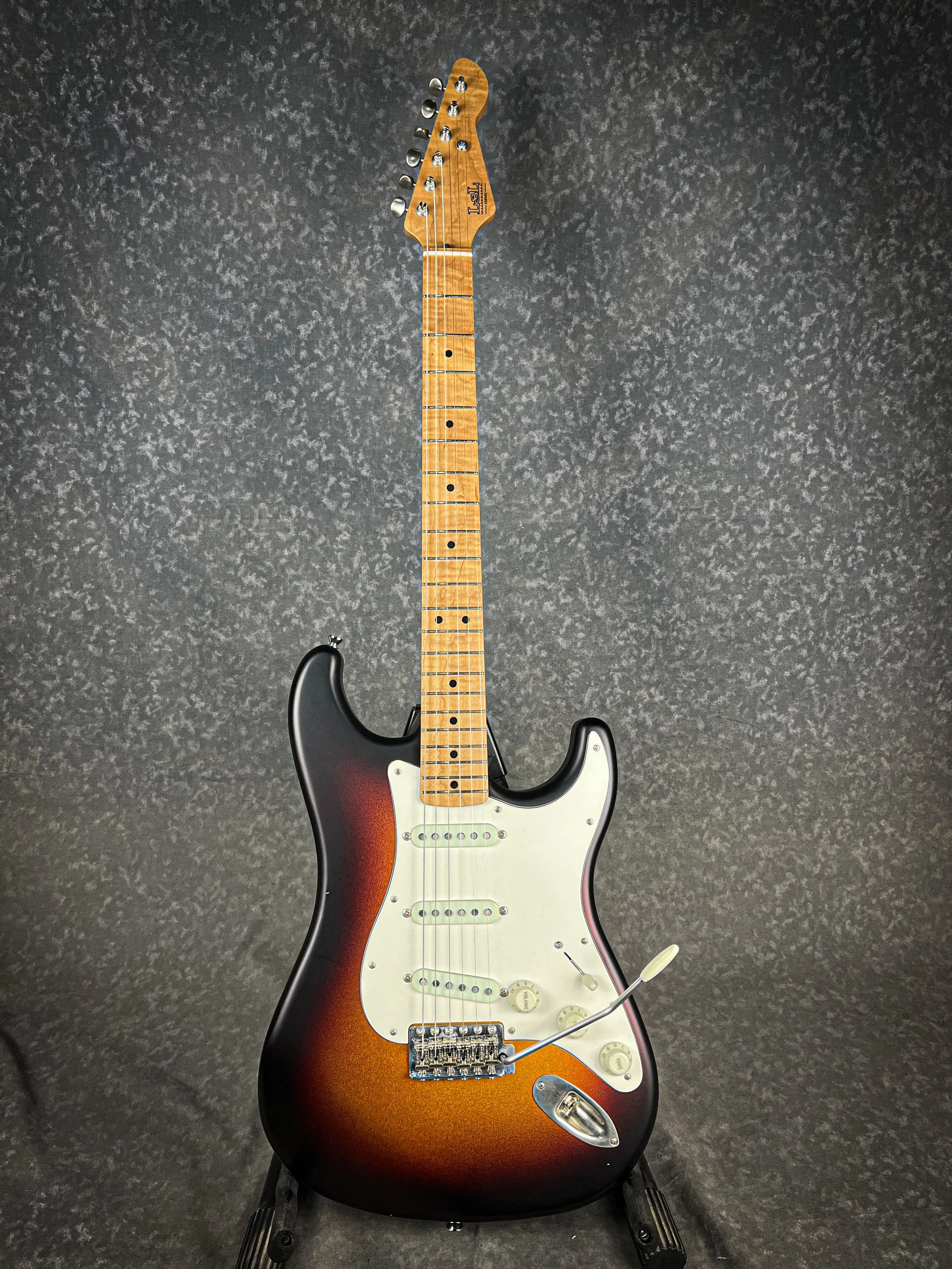 LSL Saticoy Stratocaster electric guitar with a sunburst finish and maple neck, positioned vertically on a black stand against a dark textured background.