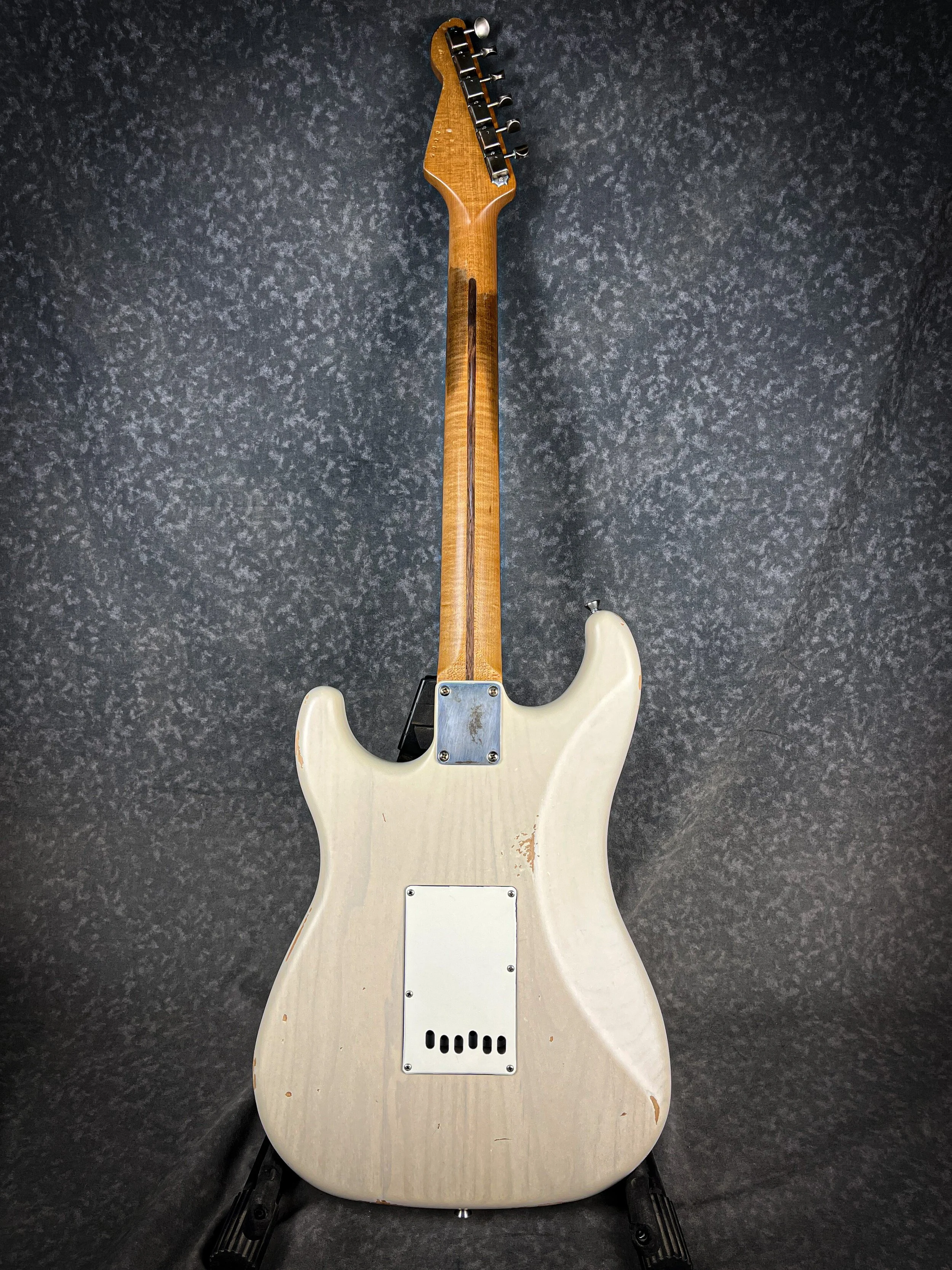 Back of a vintage LSL Saticoy electric guitar with a white worn finish, resting on black stands against a dark speckled background.