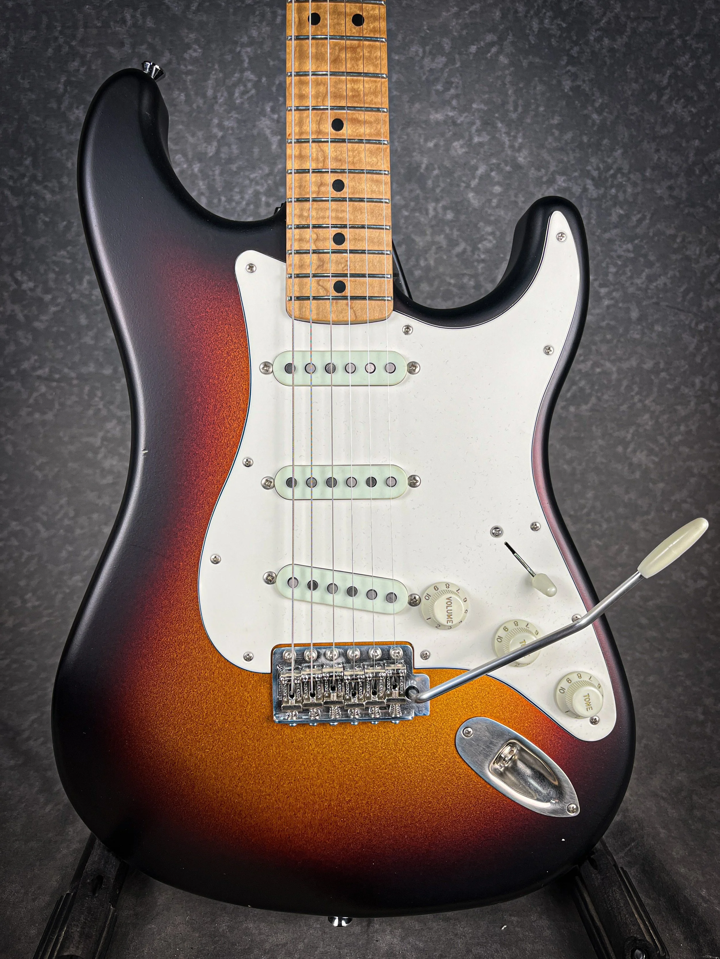 A close-up of a sunburst LSL Saticoy electric guitar with a white pickguard, three single-coil pickups, a tremolo arm, and various control knobs, set against a dark textured background.