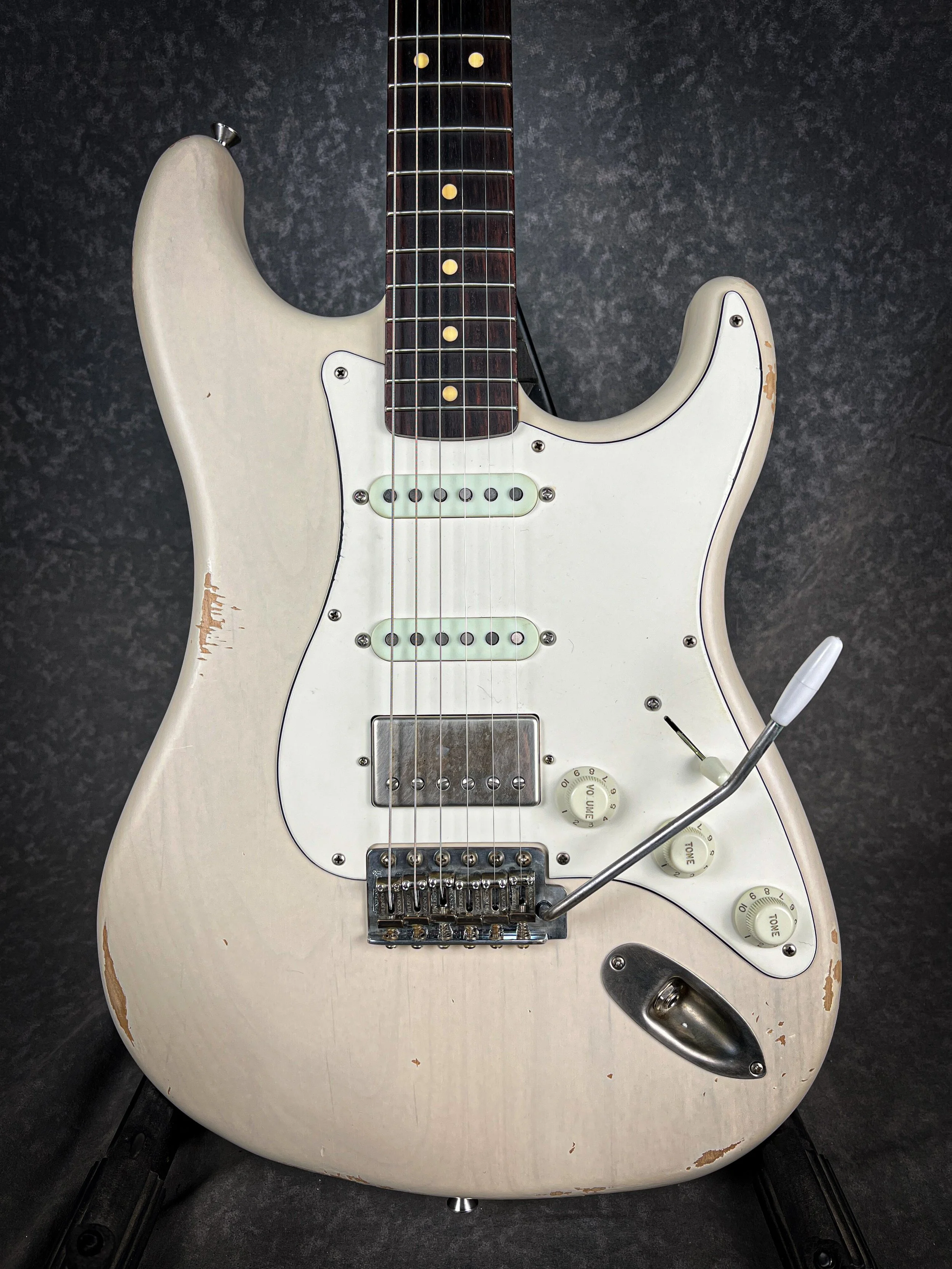 A white vintage LSL Saticoy electric guitar with a worn finish, three pickups, and a tremolo bar, set against a dark background.