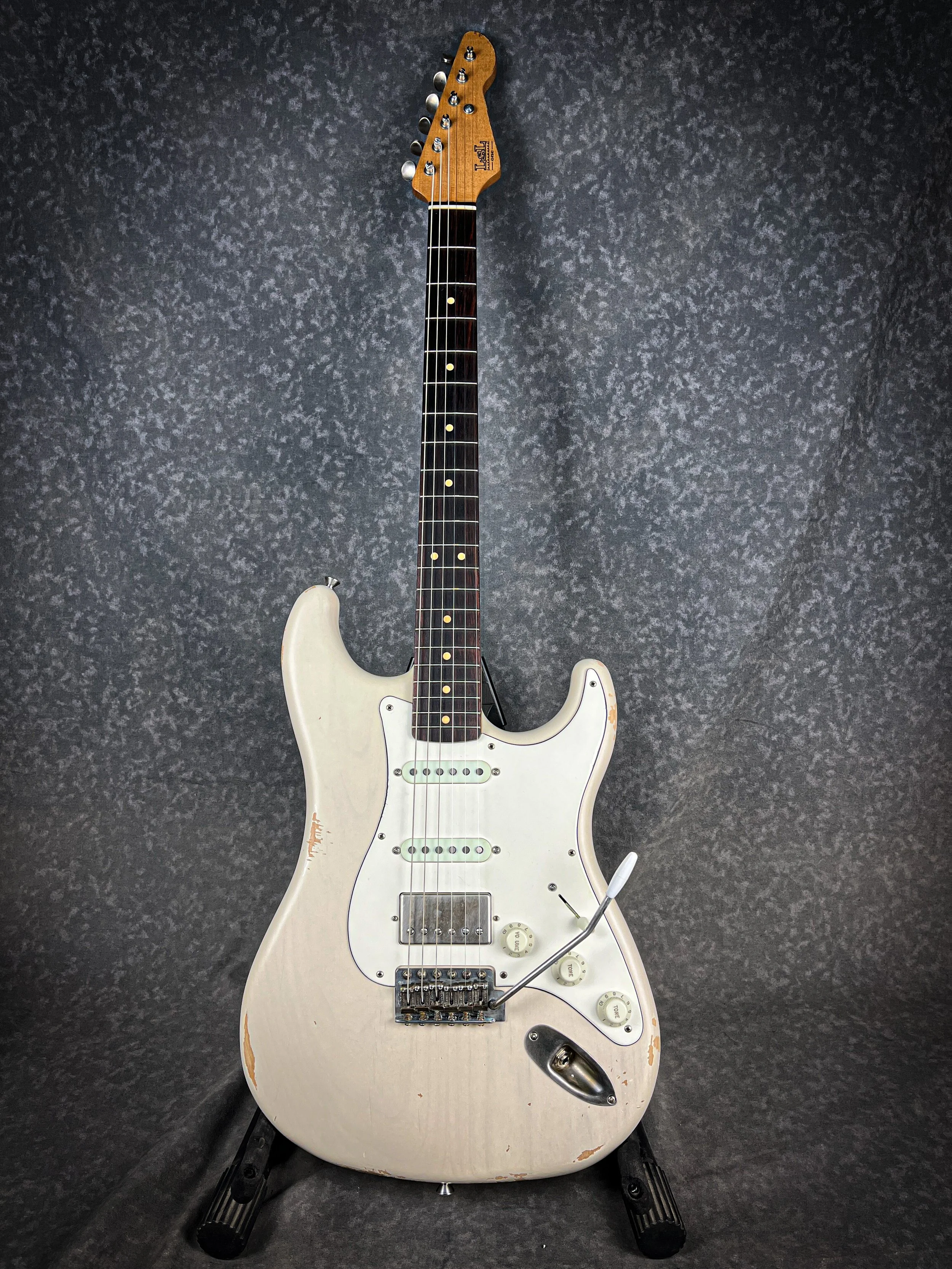 A distressed white LSL Saticoy Stratocaster electric guitar with a dark fretboard and chrome hardware, leaning against a textured gray backdrop.