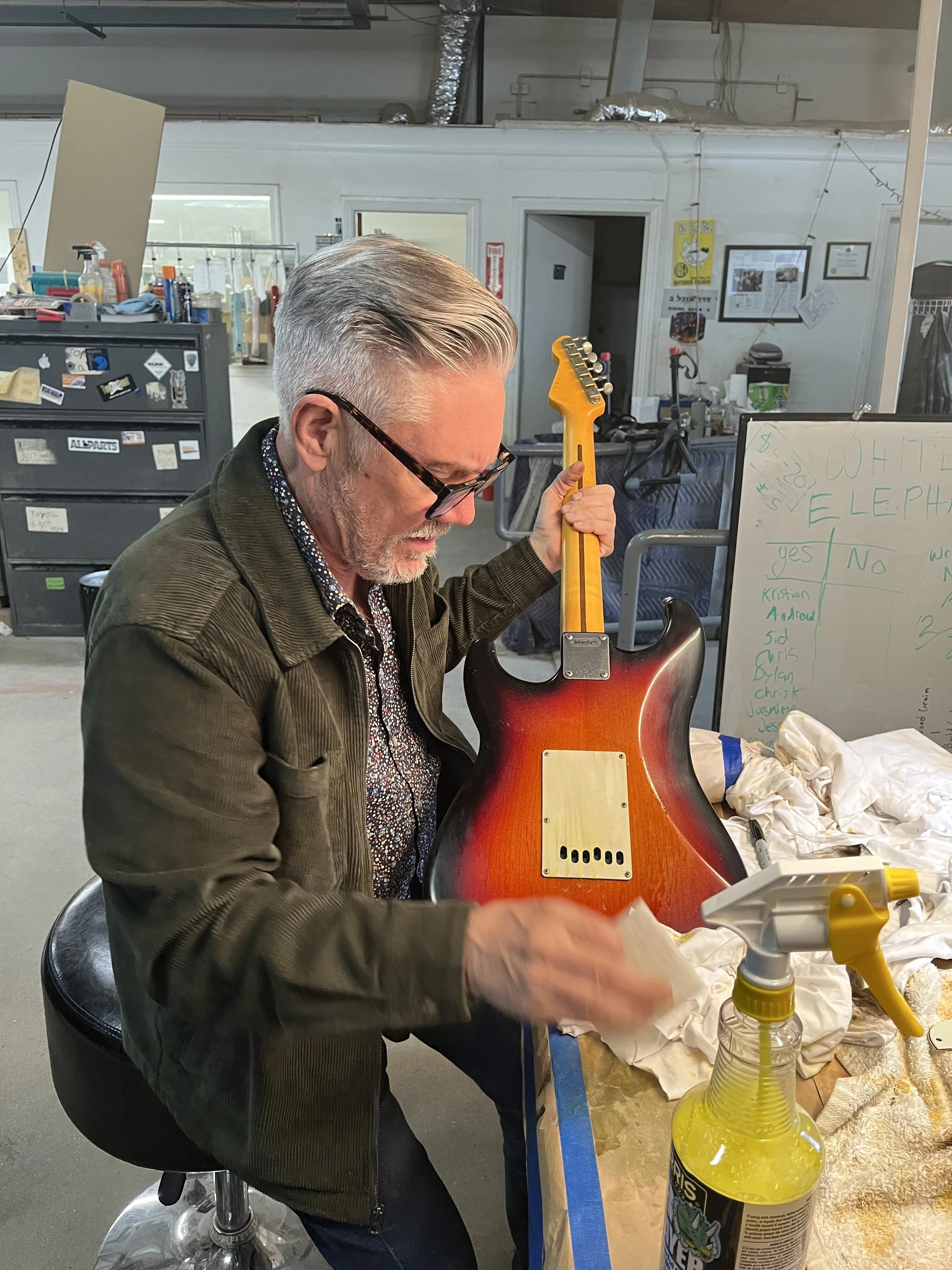 Chris Fleming working on an electric guitar in a workshop, with cleaning supplies on the table and a whiteboard with writing in the background.