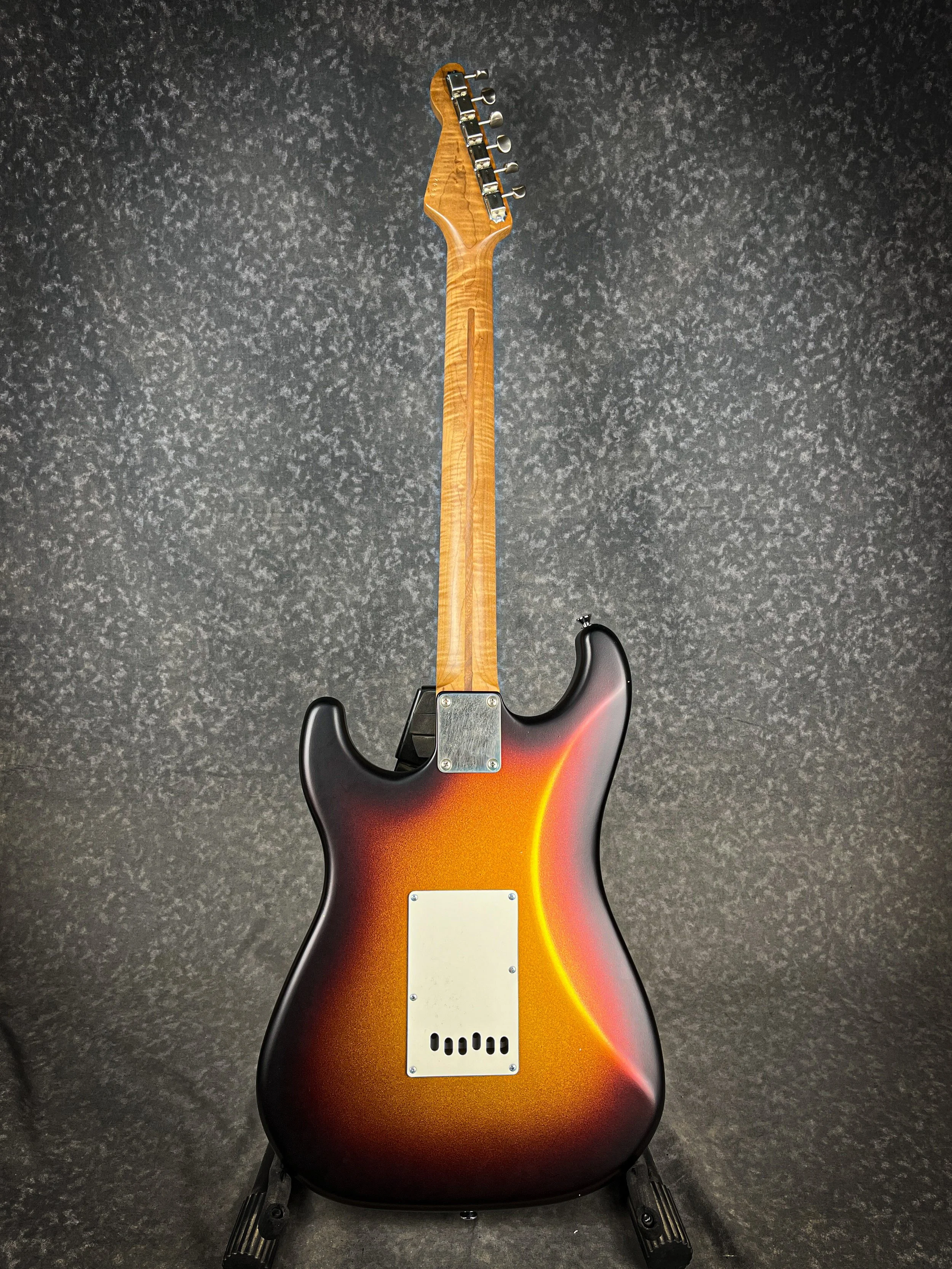 A sunburst colored LSL Saticoy electric guitar on a black stand against a dark textured background.