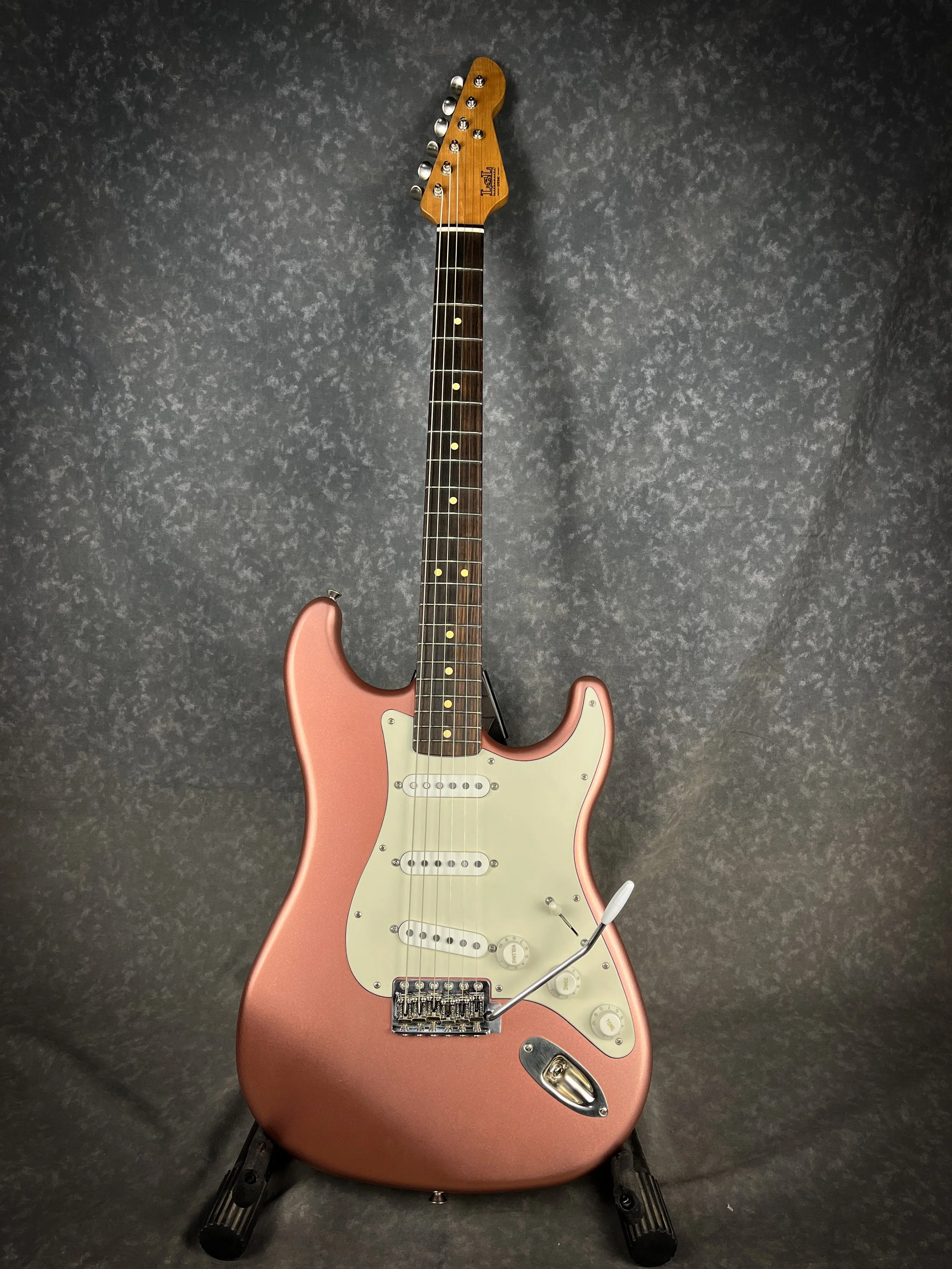 A Burgundy Mist LSL Saticoy electric guitar with a white pickguard and a whammy bar, placed on a black guitar stand against a dark, patterned background.