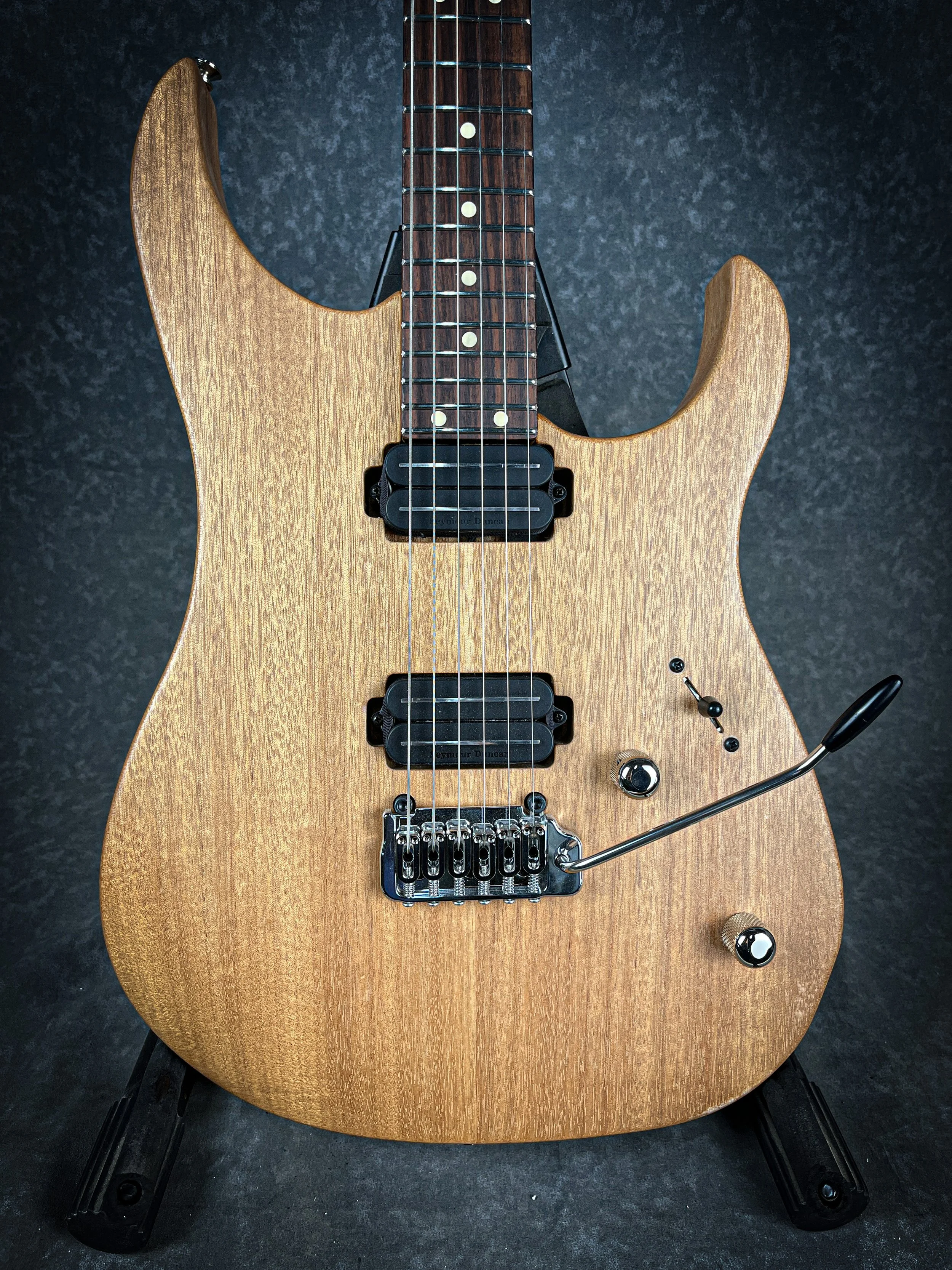 A natural wood finish LSL XT  electric guitar with two humbucker pickups, a tremolo bridge, black controls, and a toggle switch, set against a dark background.