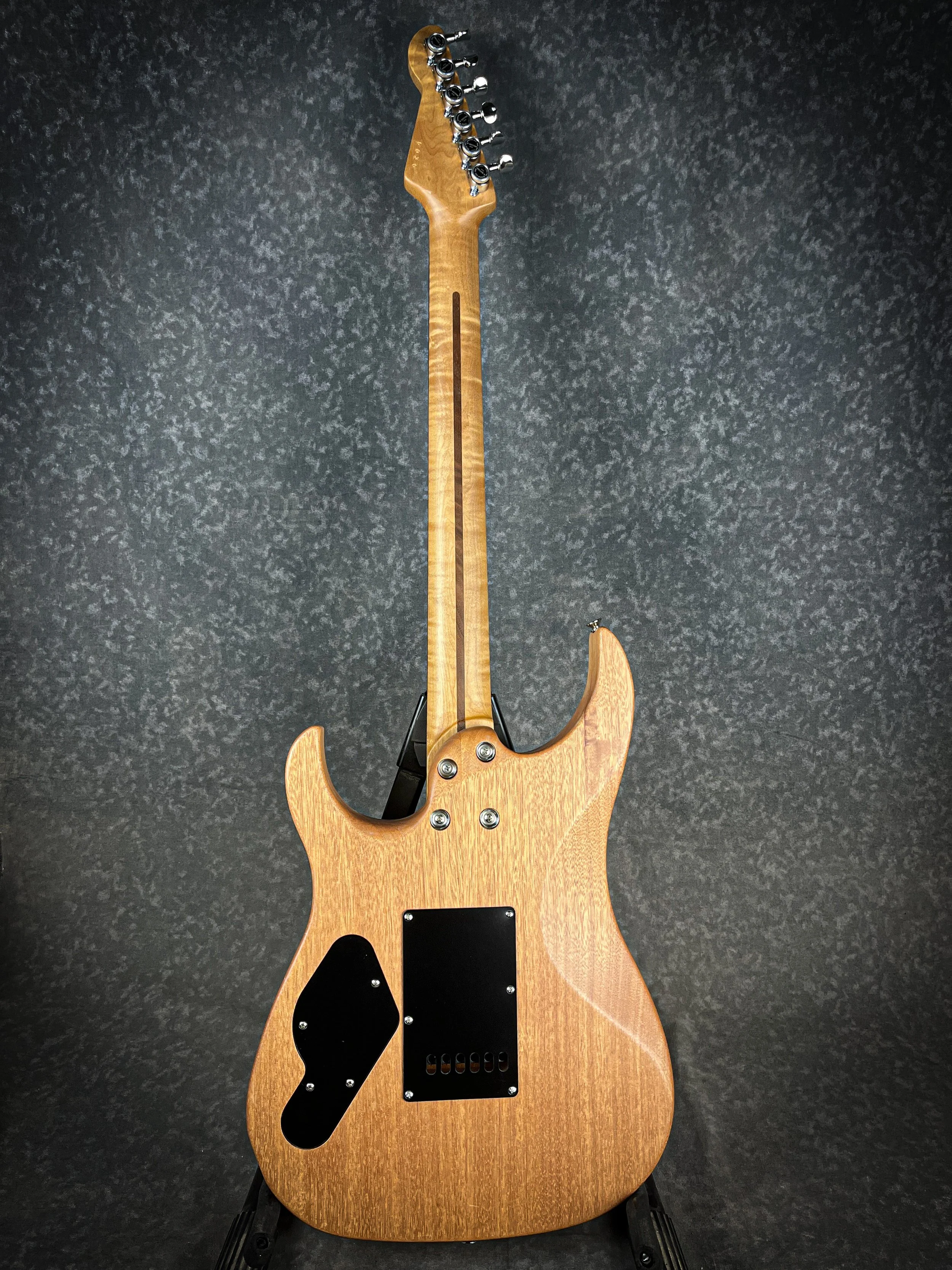 Back of an LSL XT electric guitar with a natural wood finish, black back plate, and black tuning pegs, standing on a black stand against a textured dark background.