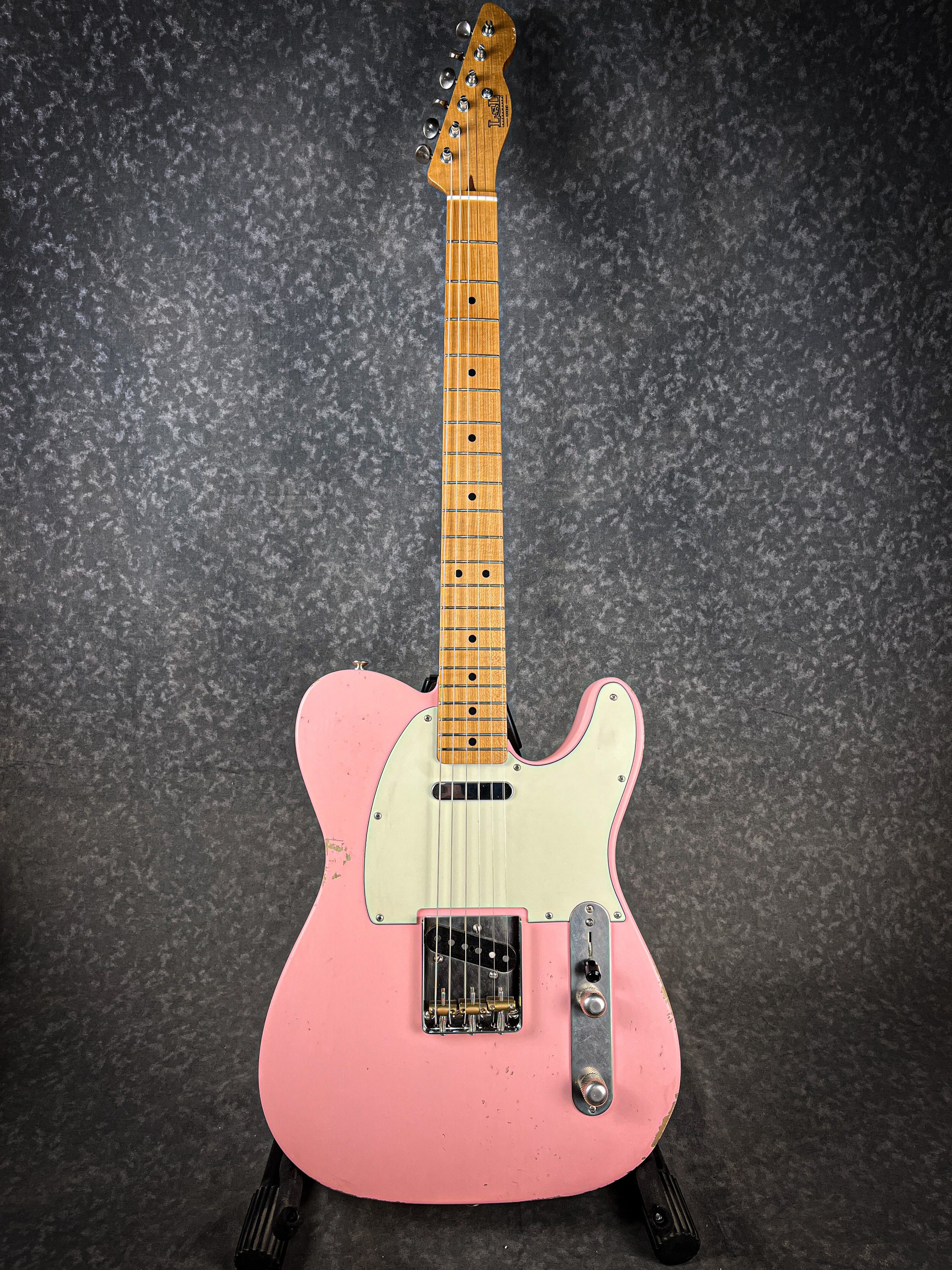 A vintage pink LSL T Bone electric guitar with a maple neck and black pickups, displayed on a black stand against a dark textured background.
