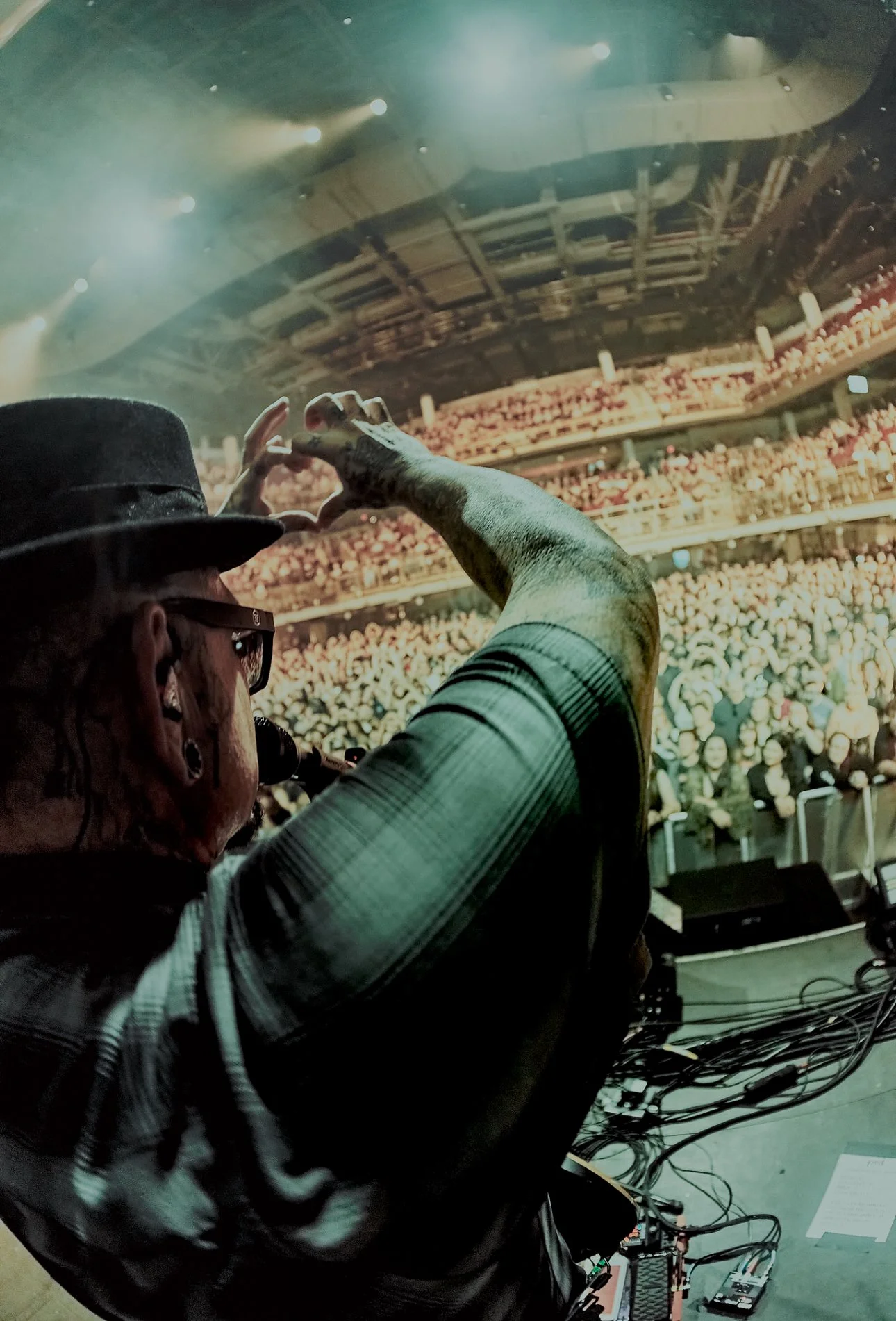 Marcos Curiel wearing glasses and a hat, making a heart shape with his hands while performing on stage at a concert with an audience in the background.