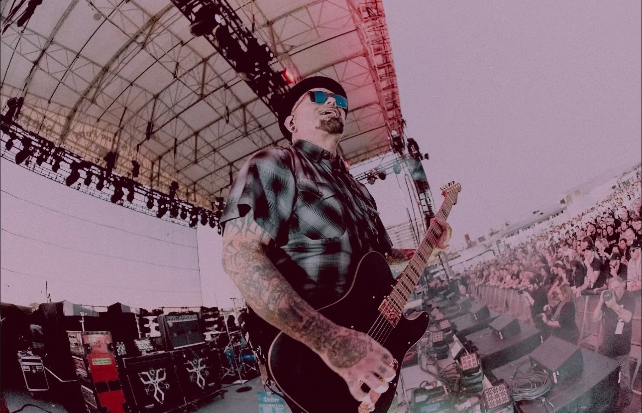 Marcos Curiel wearing sunglasses, a plaid shirt, and a black hat playing an LSL Instruments boutique electric guitar on an outdoor stage during a concert, with a large audience watching.