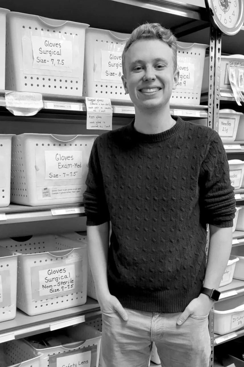 A young man with short light hair, smiling and standing with hands in pockets, in front of shelves filled with labeled plastic bins containing medical supplies such as gloves, eye protection, and catheter accessories, in a storage or supply room.
