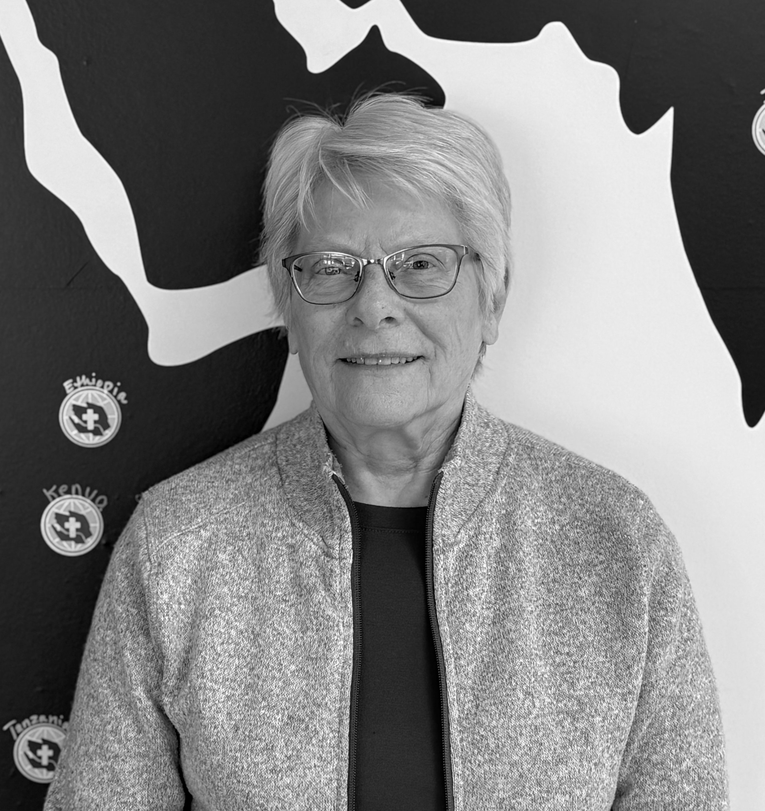 A smiling elderly woman with short gray hair and glasses, wearing a dark shirt.