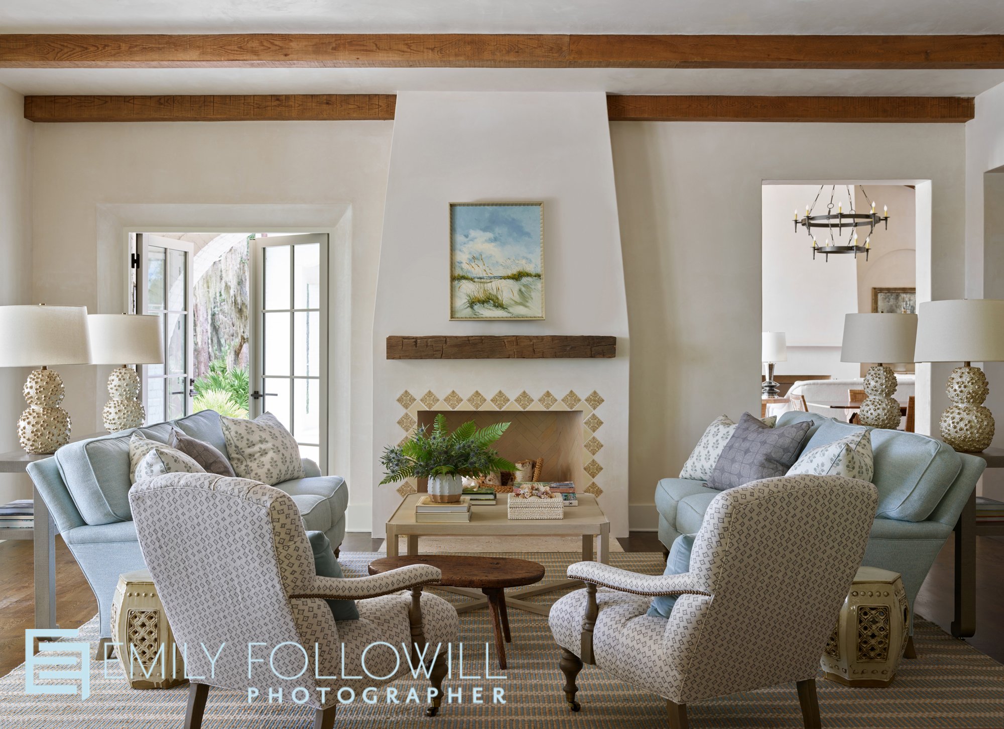 small living room with blue and white furniture. Wooden beams stand out in the ceiling and above the un lit fire place. The doors are open revealing the live oak trees on this Sea Island Georgia home.  Designed by Courtney Giles and published in Atla