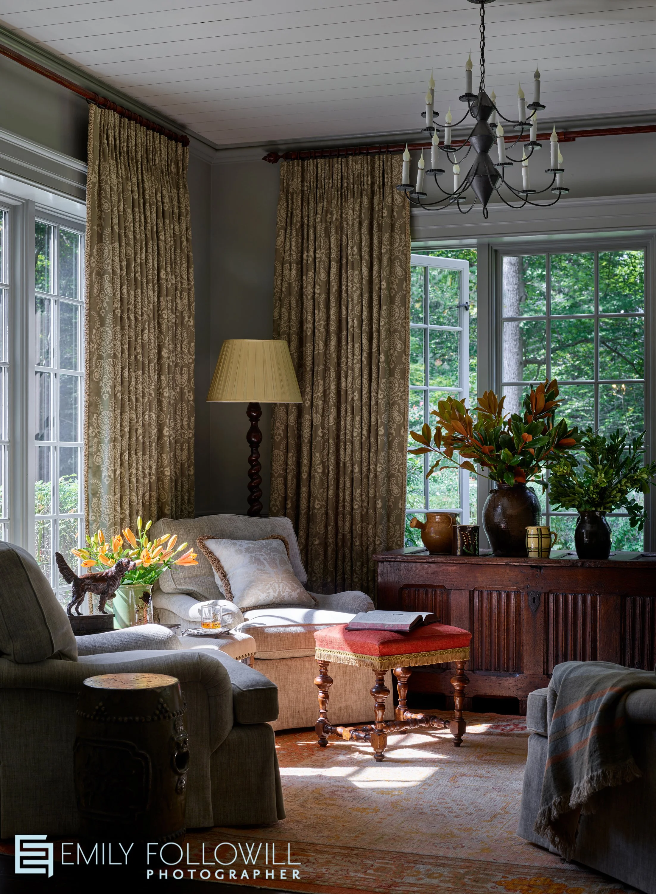 moody grey sunroom with light spilling onto the orange flowers on the side table. Patterned drapes with a hint of yellow hanging on the opened windows. Magnolia sits on the large side board. Designed by Jackye Lanhem in Atlanta Georgia 