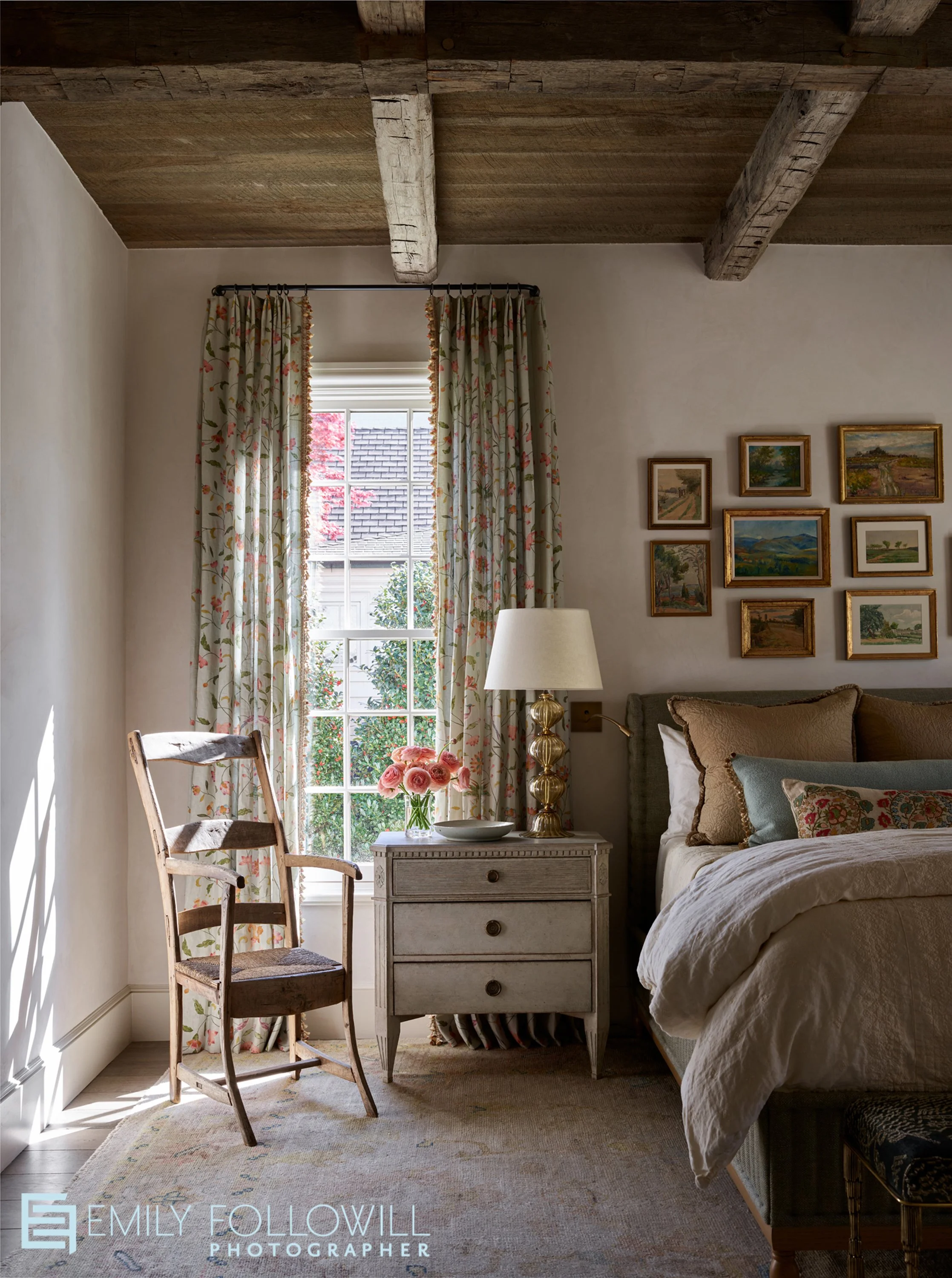 A creamy walled moody bedroom with rustic wooden beams above the cozy bed. A rustic wooden chair sits next to the bedside table. Sunlight streams through the window. Location: Atlanta, Georgia