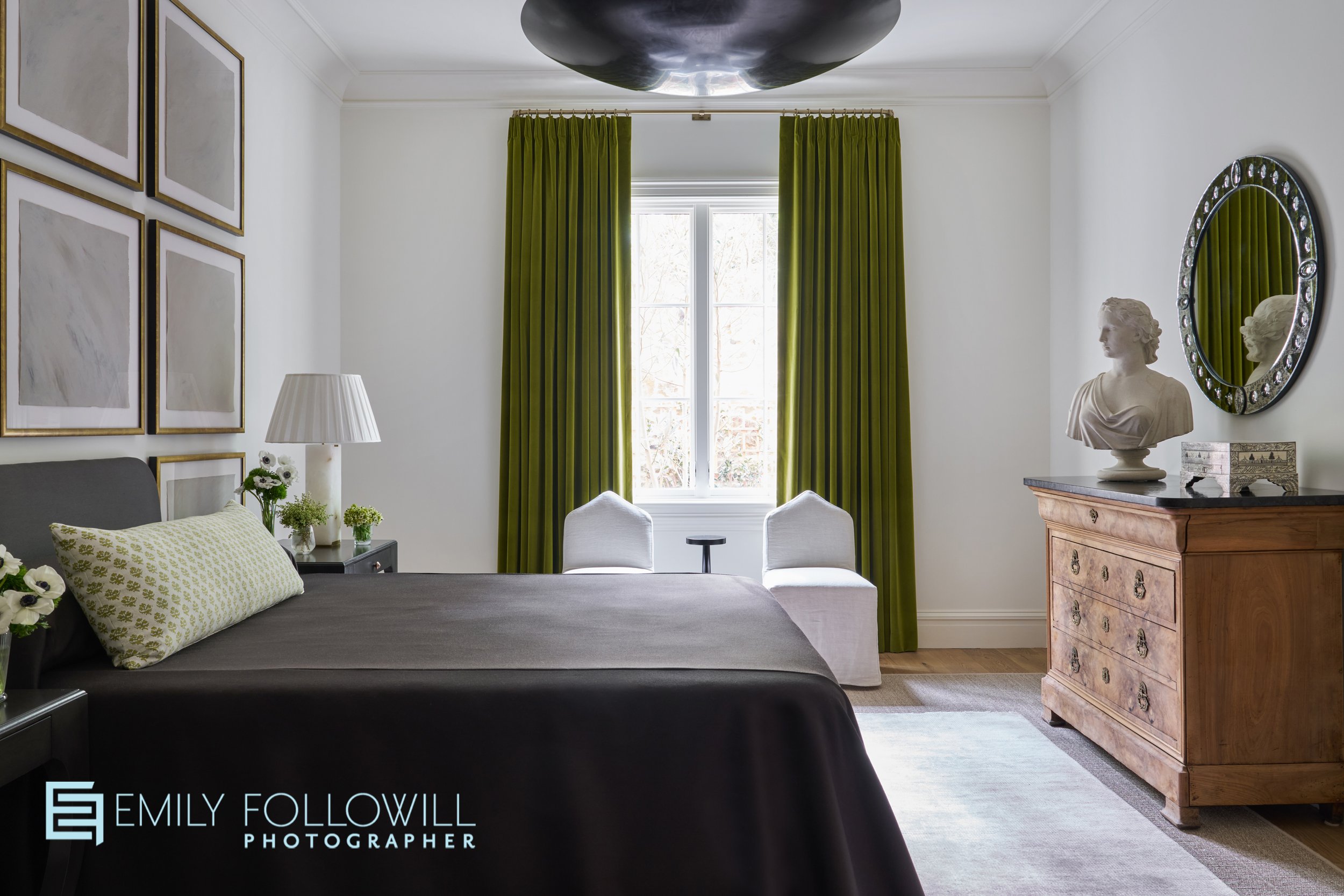 Primary bedroom wigh white walls, green drapery, black bedding, and custom art work above the bed. A statue of a woman sits atop of an antique dresser by a round mirror. a large round black chandelier hangs in the middle of the room. Location, Atlant