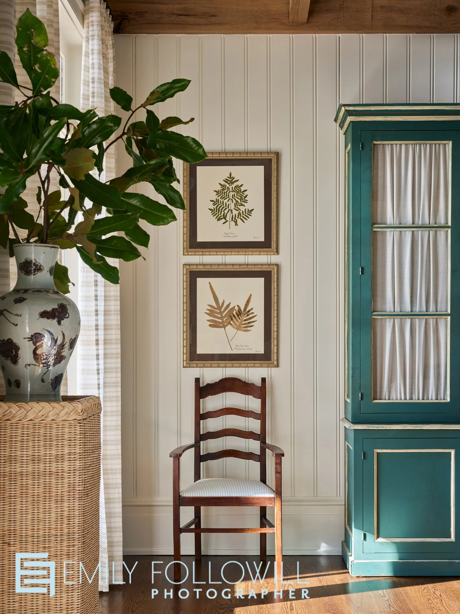 detail of a green wooden cabinet, with a rustic chair underneath two floral prints. Large magnolia in an urn int he foreground. Light spilling in from the draped windows. Located in Fairhope Alabama. Designed by Ashley Gilbreath