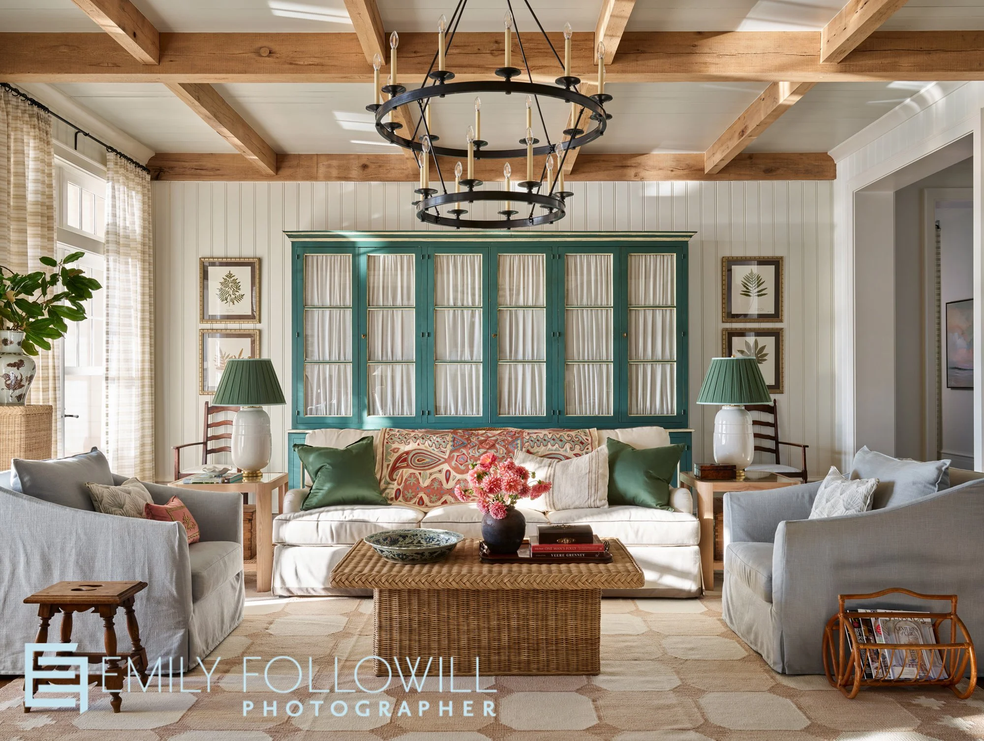 Large living room with sunlight streaming through. White linen furniture, with hand painted green cabinetry. A wicker coffee table ties in the wooden beams on the ceiling. Location, Fairhope Alabama. Designed by Ashley Gilbreath. 