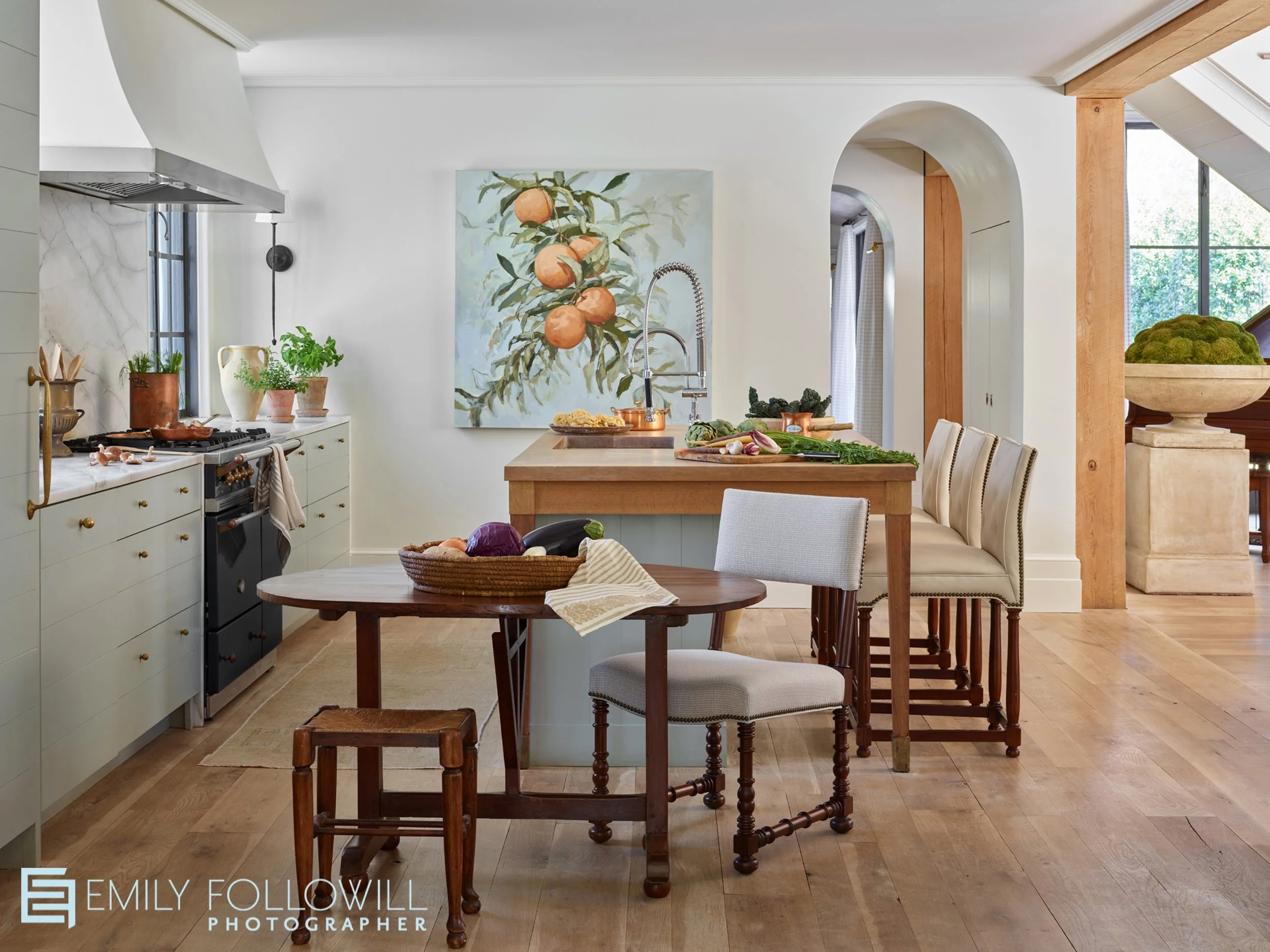 white kitchen with an arched doorway to the hallway. A rustic wooden table sits in front of a wooden island. A large painting of peaches hangs on the wall behind the kitchen island. Design by Ashley Gilbreath. Location: Alabama