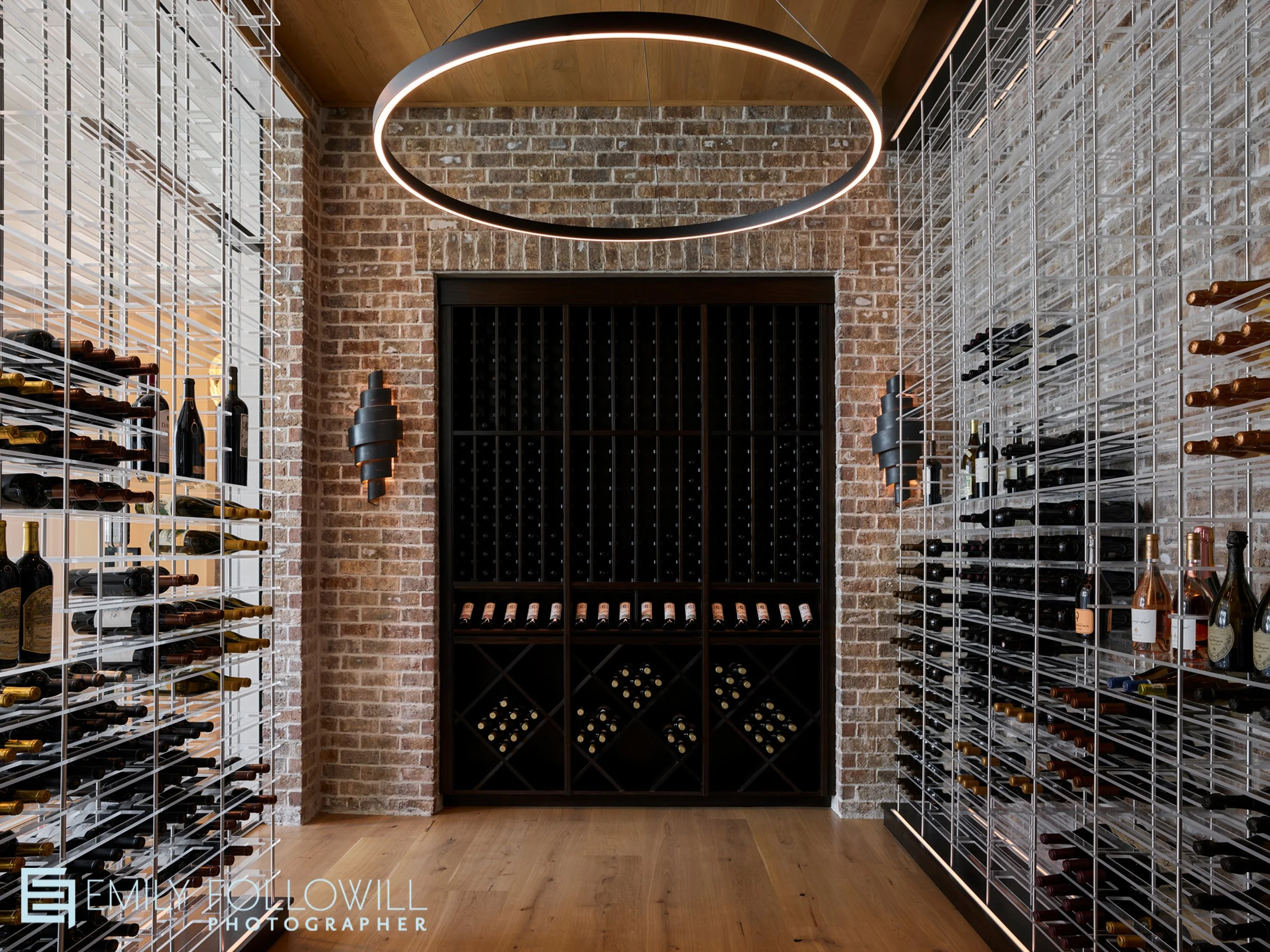 sleek modern wine cellar surrounded by glass walls looking into the brick wall. A large circular chandelier is illuminated above deep mahogony shelves for the wine. Located in Sea Island Georgia. Designed by Abigail Interiors