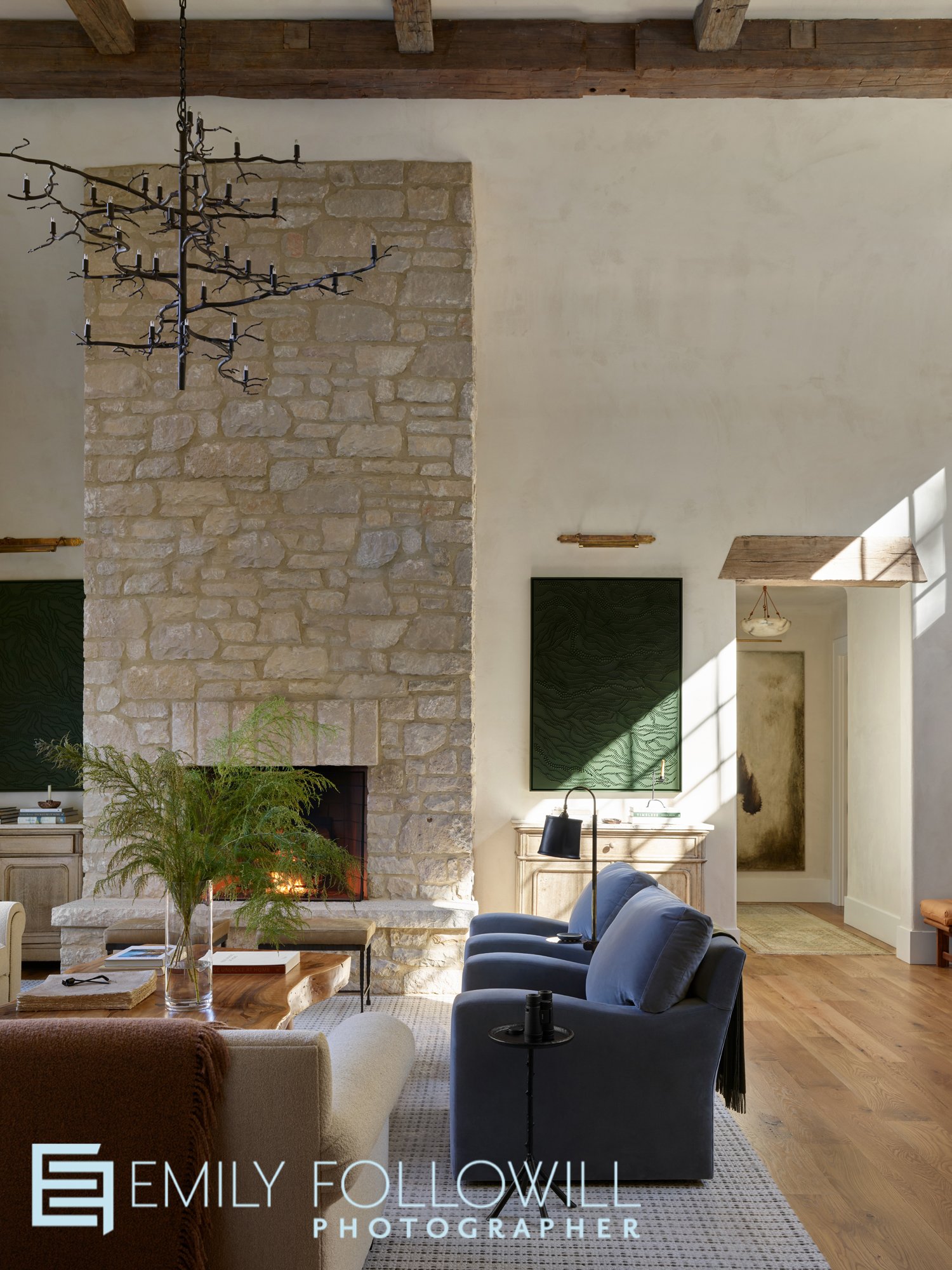 Lofted wooden beamed ceilings soar above this sunlit living room. A large stone fireplace invites you to have a seat in one of the blue chairs and take in the views of Lake Burton. Designed by Melanie Milner