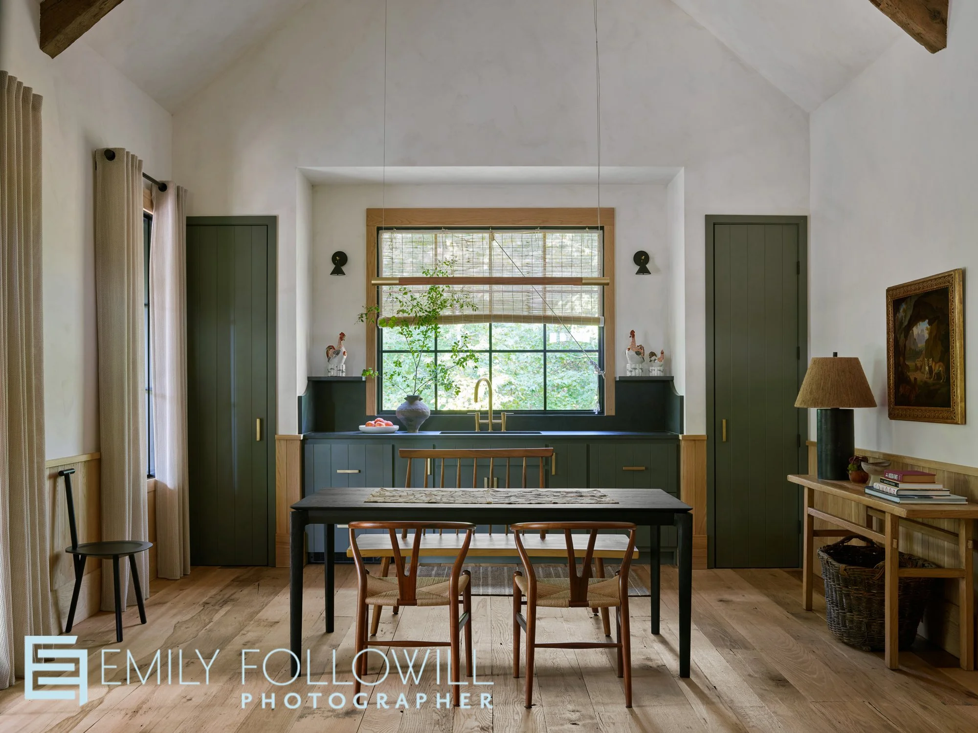 Large open kitchen with vaulted ceilings, and green cabinetry. The sink sits below a large window overlooking lush greener. A large wooden dining table awaits its guests
