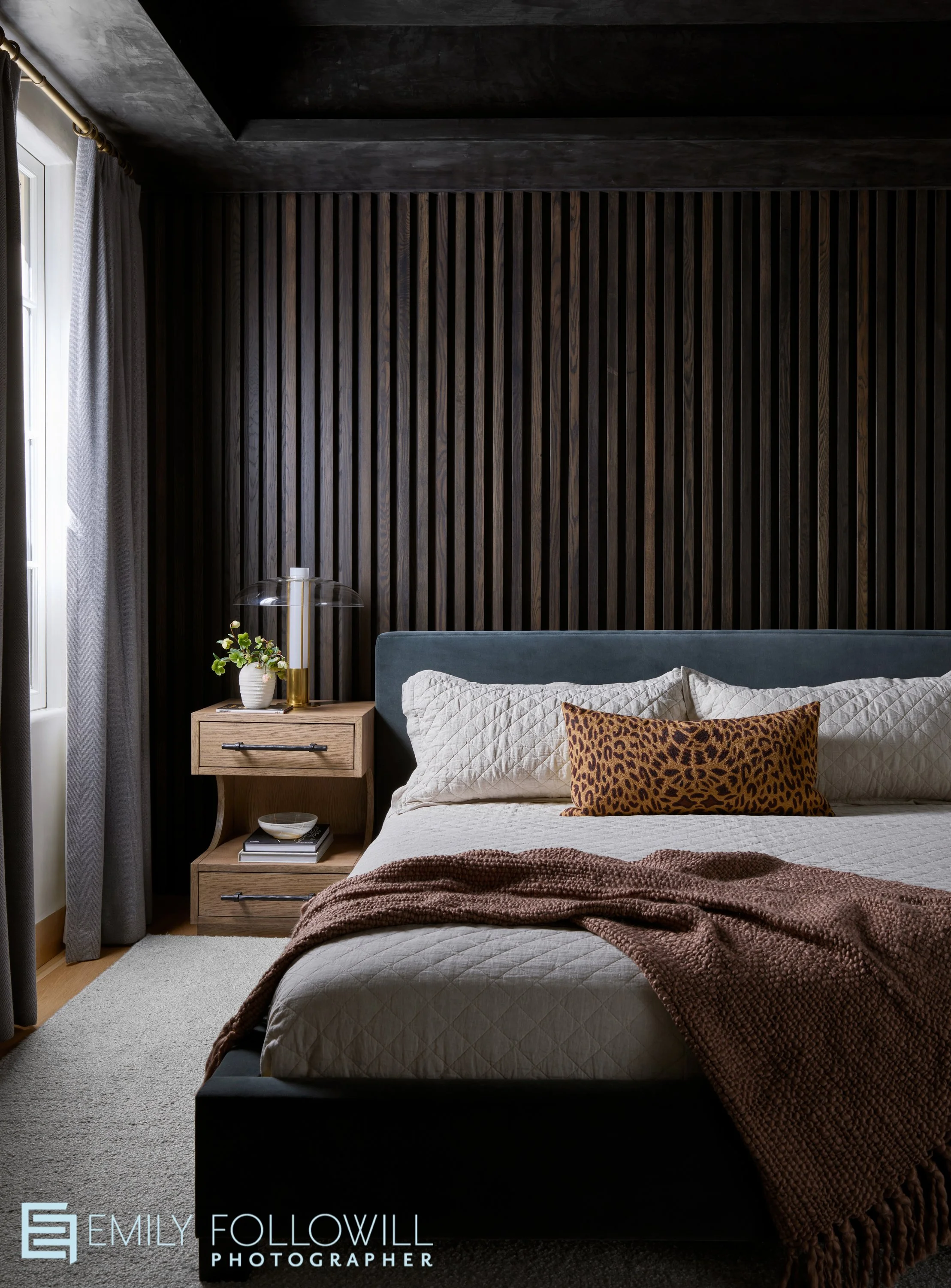 Moody bedroom with wood paneled walls. Natural light softly streams across the wooden bedside table and blue velvet bed frame.Designed by Nancy Duffey Interiors