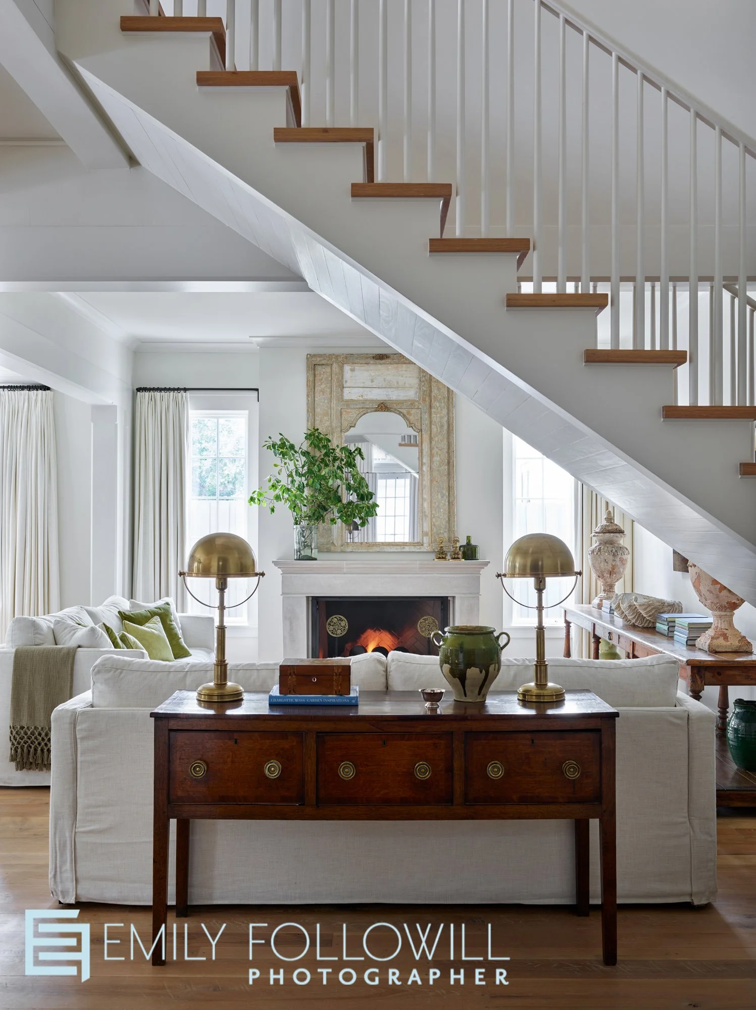 A bright white living room sits behind the staircase to upstairs. Gold lamps sit upon the antique side board behind the white linen sofa. Location: Rosemary Beach Florida Architect: TSB Architecture