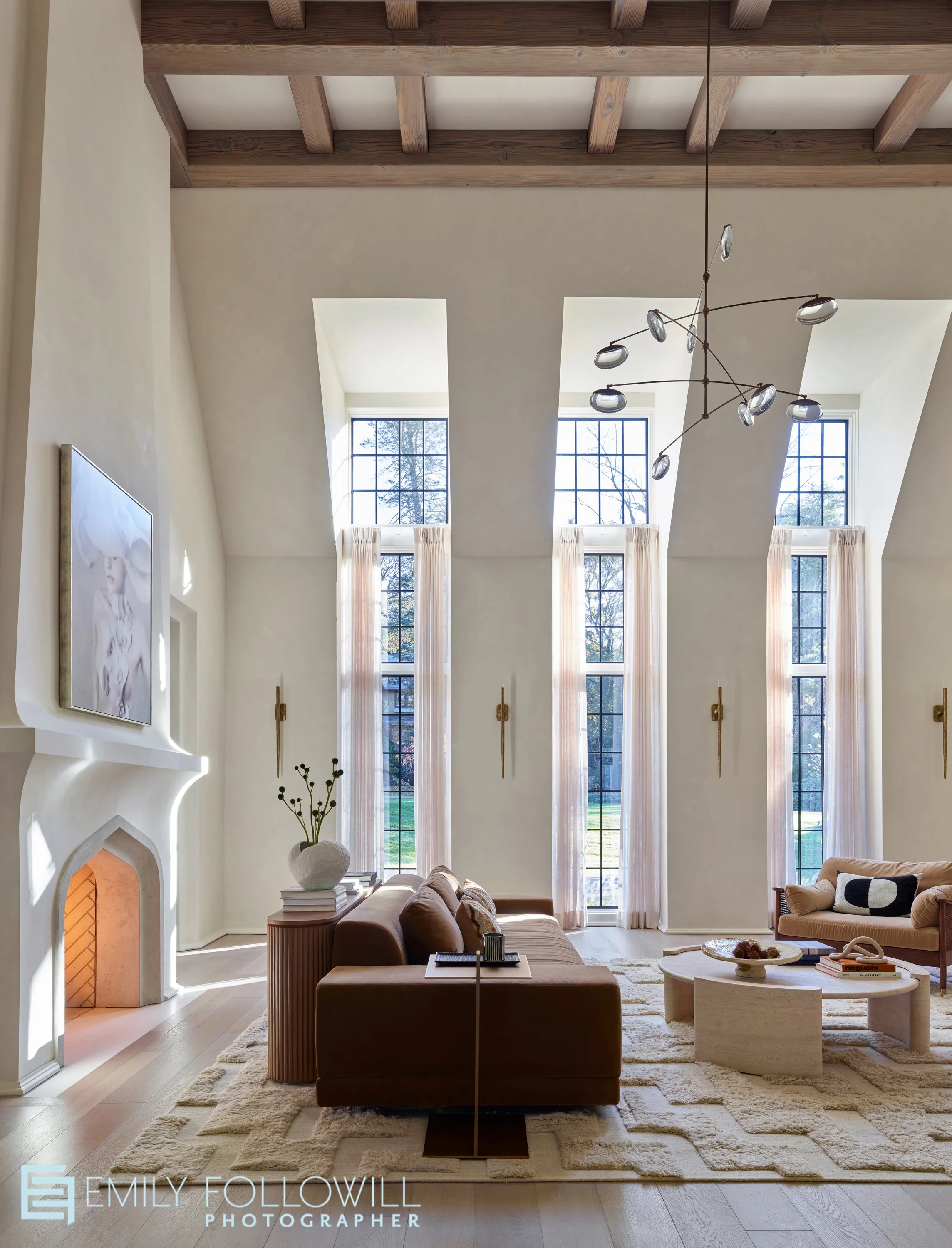 Large living room with a stone fireplace and three tall windows. Organic modern furniture illuminated by the warm natural light. Located in Rochester New York. Designed by Christopher AI
