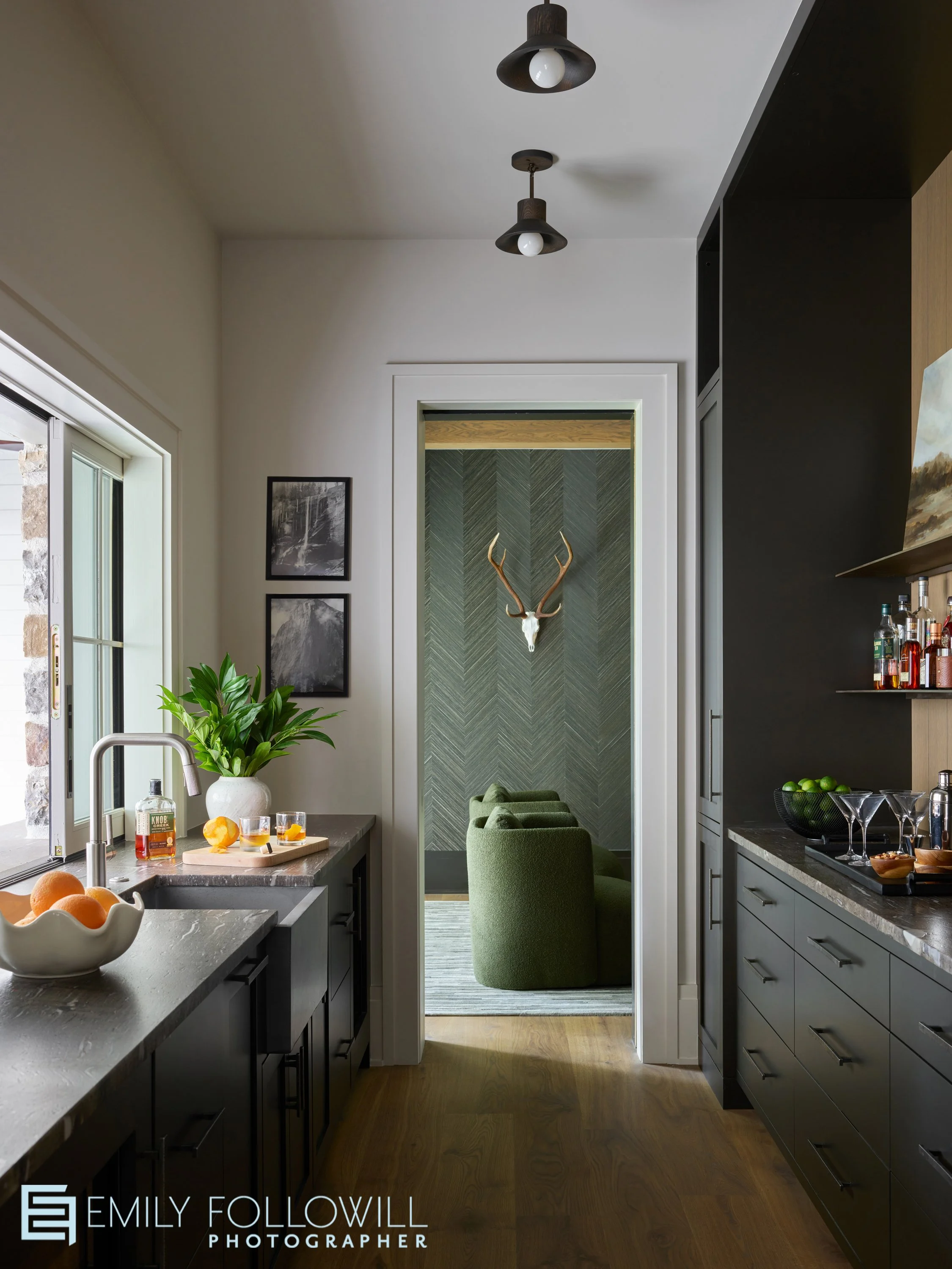 sleek bar or butlers pantry looking into the green room beyond. deer antlers hang above two green chairs. Nano windows above the sink are open.