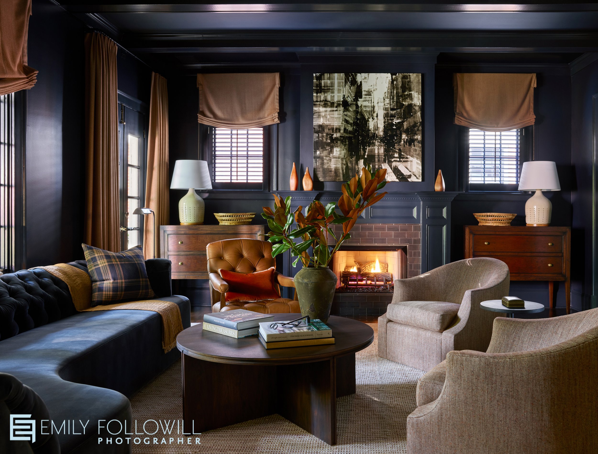 Moody formal living room with deep blue walls, modern velvet sofa, a cozy lit fireplace with two white chairs flanking the rich wooden coffee table. Located in Ansley Park, Atlanta Ga. Designed by Melanie Milner