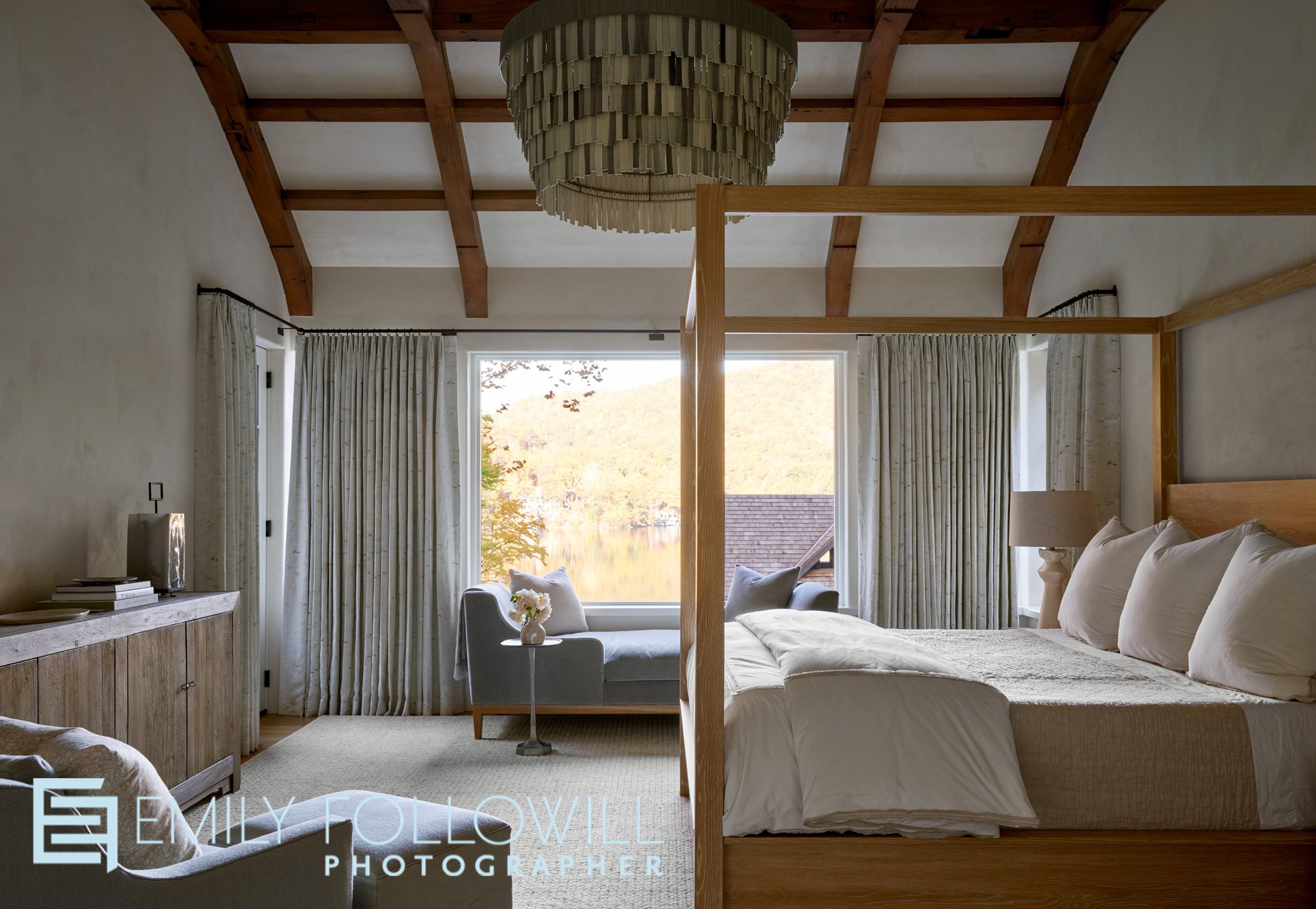 Venetian Plaster walls surround this serene primary bedroom overlooking Lake Burton. Soft bedding, and  a chaise lounge to have a cup of tea and read while on vacation, creates a peaceful scene. Designed by Melanie Milner