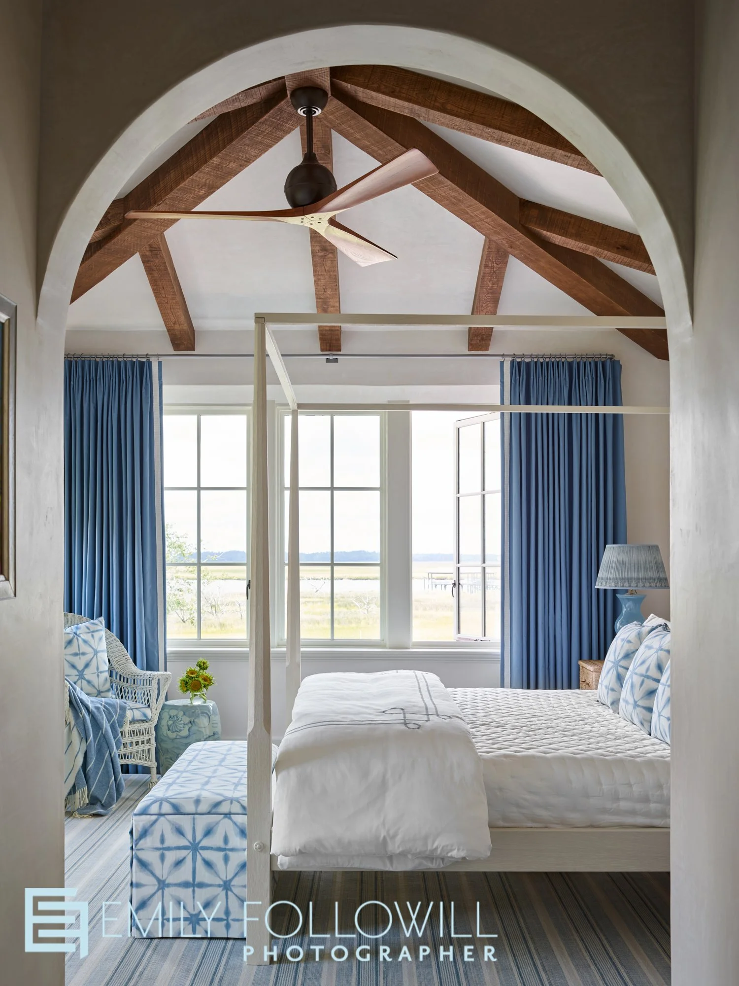 Bright happy guest bedroom seen through an arched entry-way. Wood beamed ceiling above the four poster bed. Large windows with blue drapery provide a view to the ocean in the Sea Island Georgia home. Deisgn by Courtney Giles. Published in Atlanta Hom