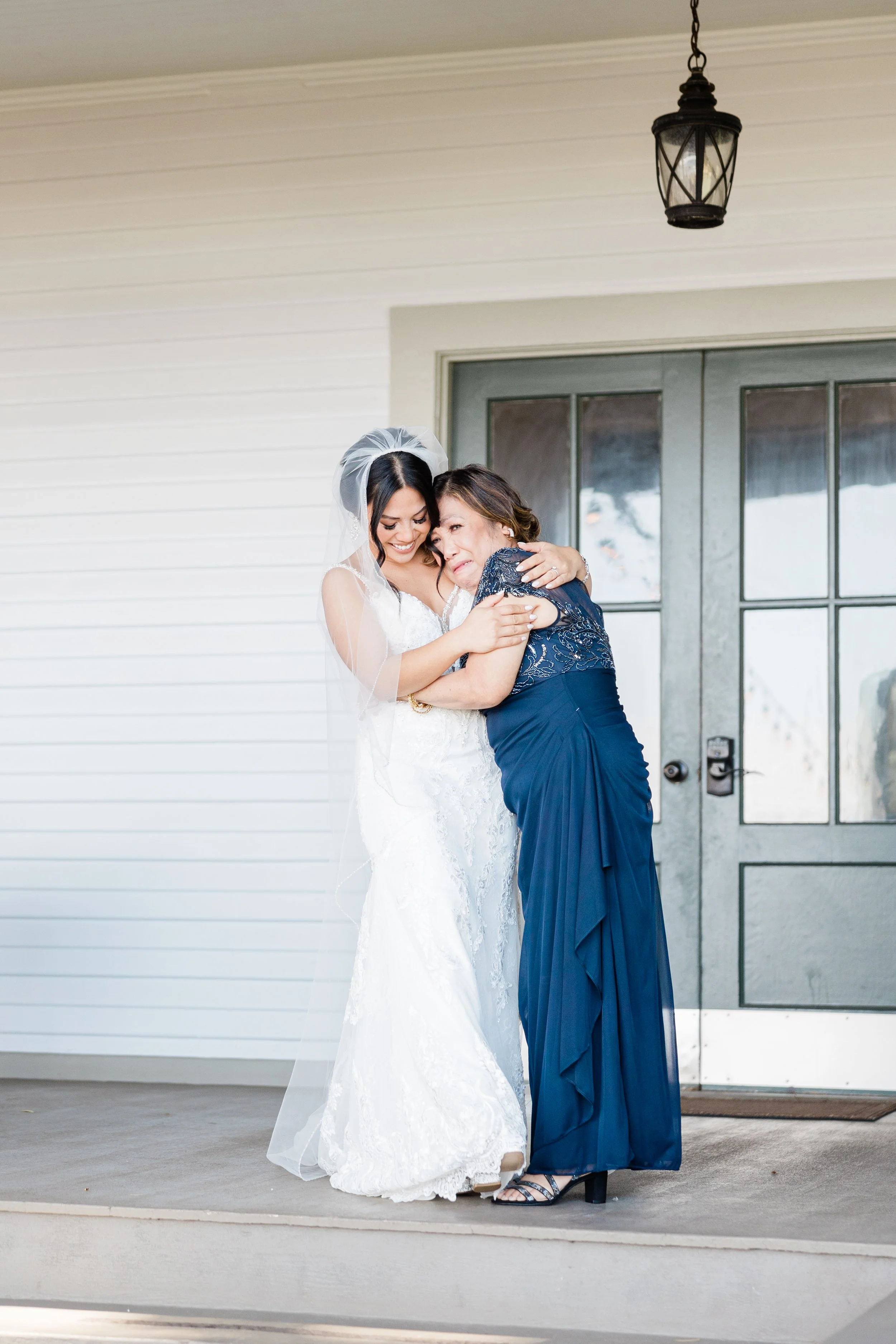 A bride in a white lace wedding gown and veil is hugging an older woman in a navy blue dress outside a building with a large gray door and white siding, under a hanging black lantern.