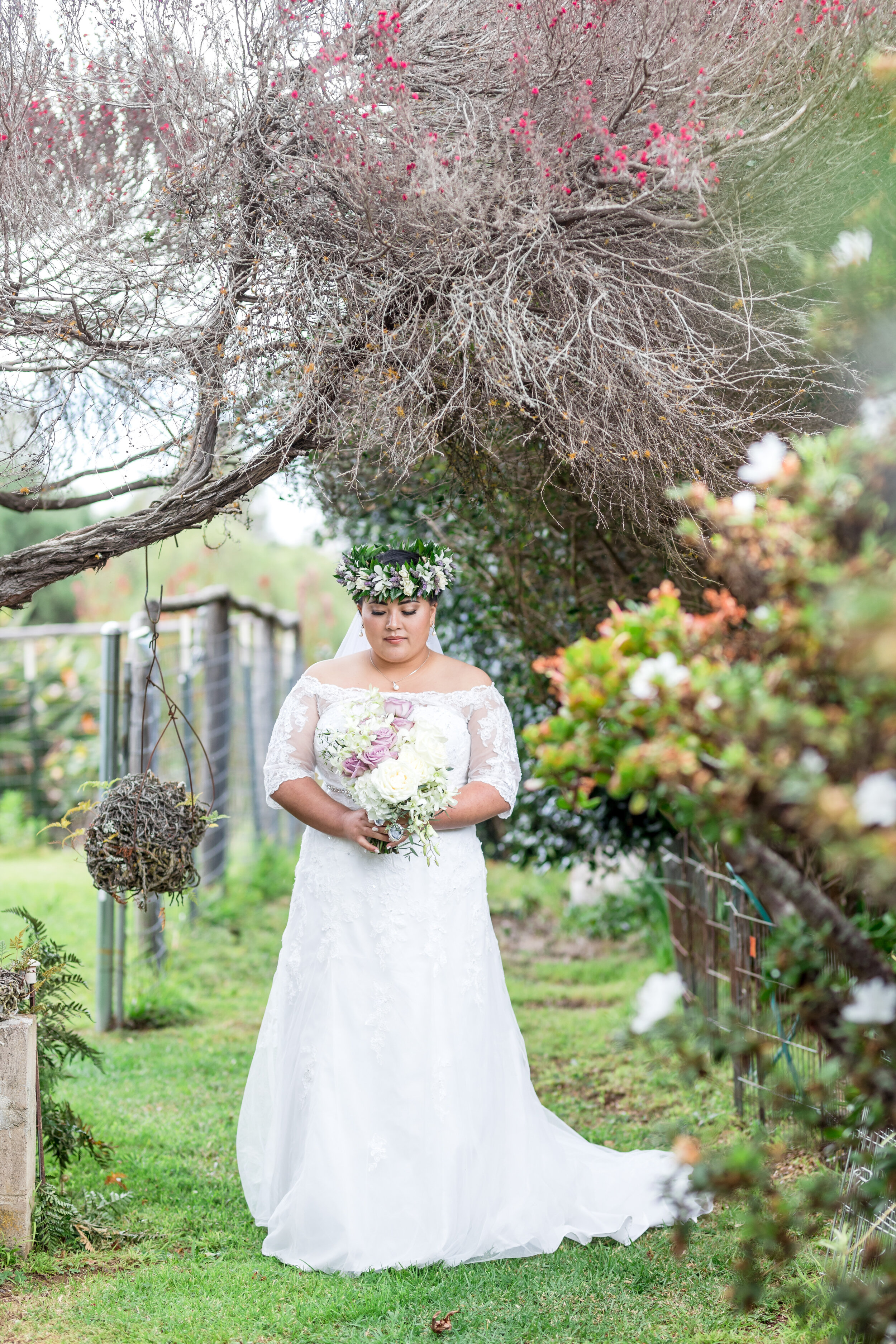 A bride in a white wedding dress holding a bouquet of pink and white flowers, standing in a garden under a large, leafless tree with pink blossoms.