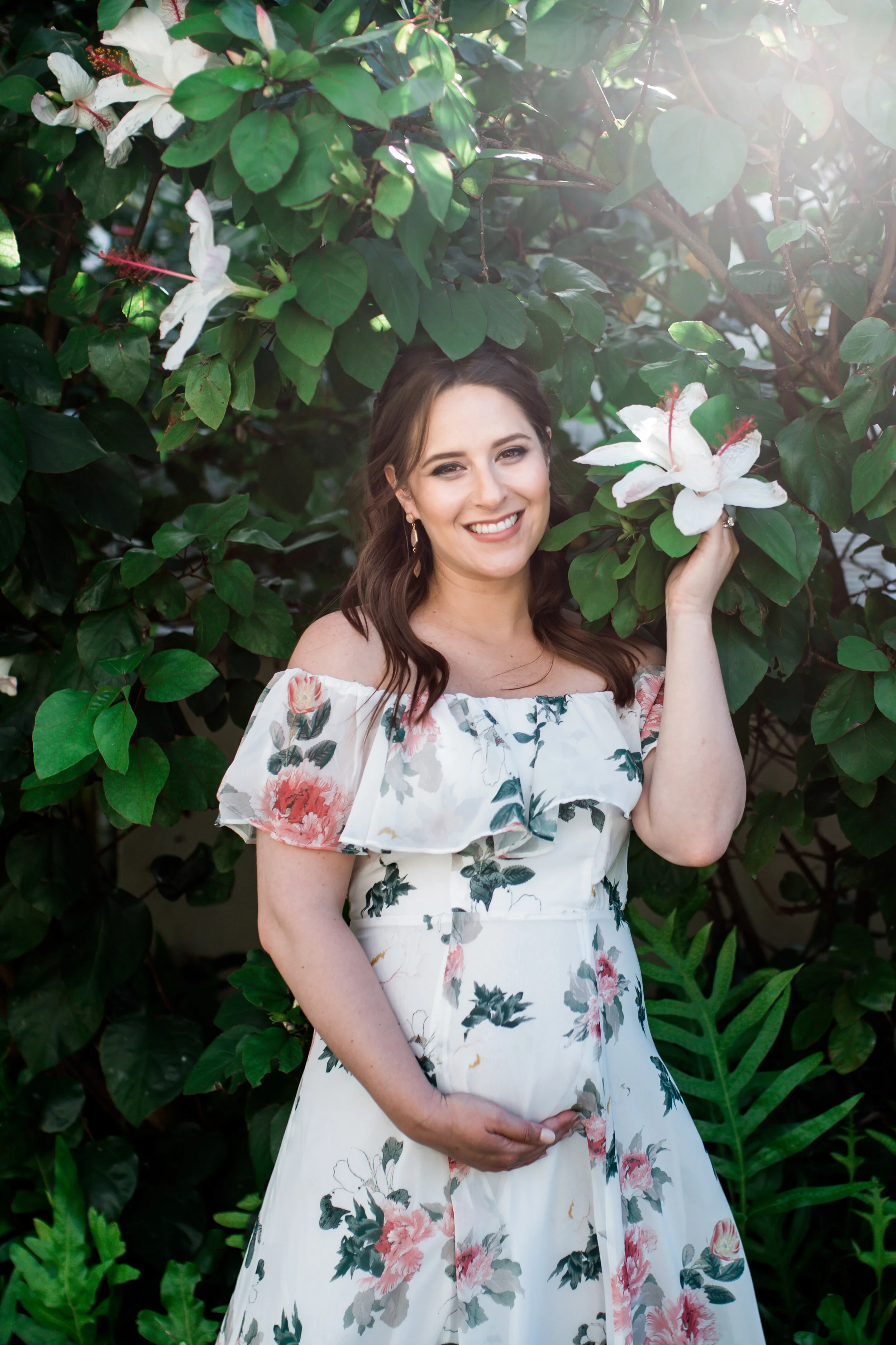 A woman with brown hair wearing a floral off-shoulder dress, smiling, standing among green leaves and white flowers, holding a flower in her right hand.