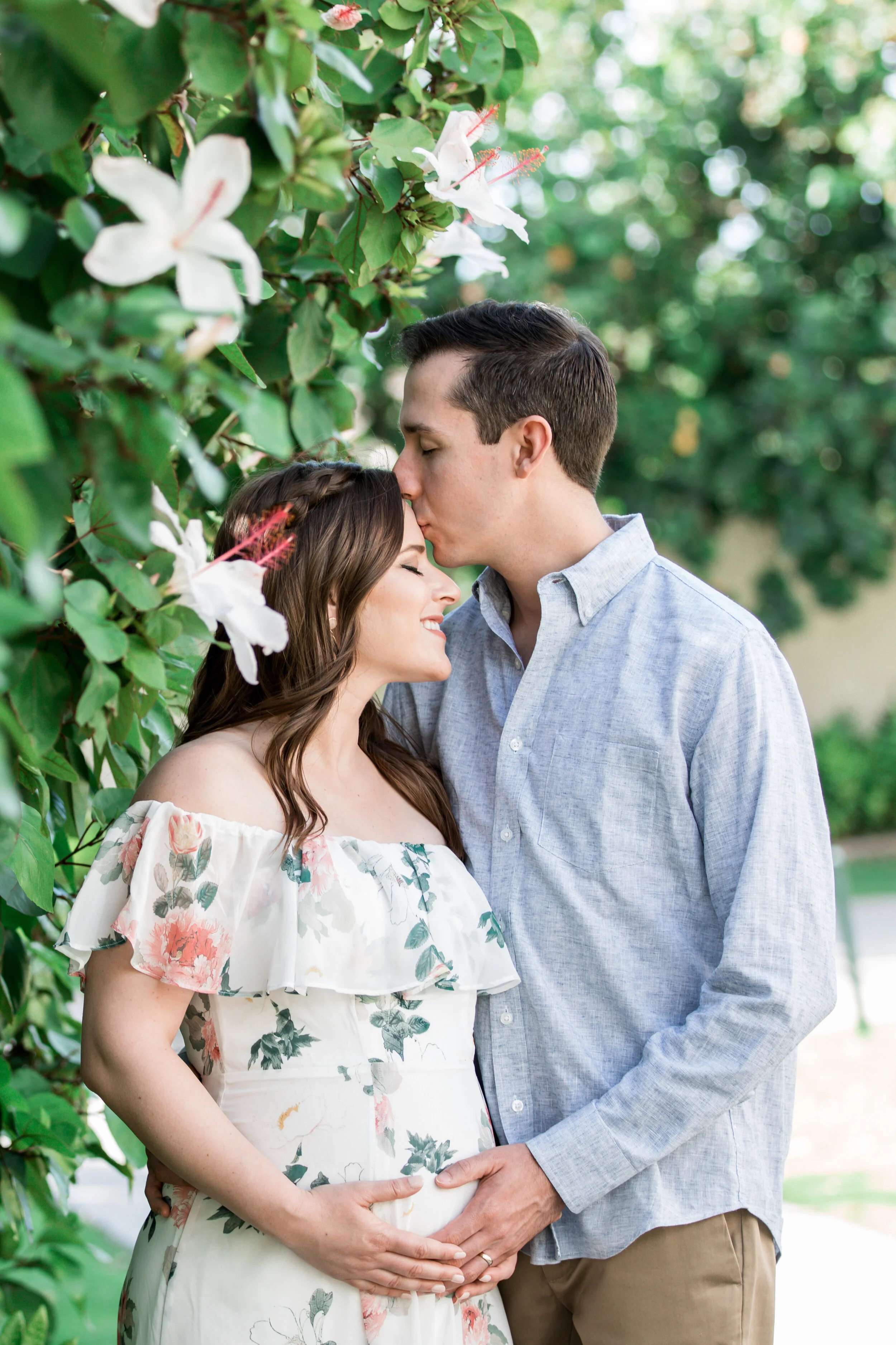 A pregnant woman with long brown hair, wearing a floral off-the-shoulder dress, kissing a man with short dark hair, wearing a light blue button-up shirt, as they stand outdoors amidst greenery and white flowers. The man is holding the woman's baby bu