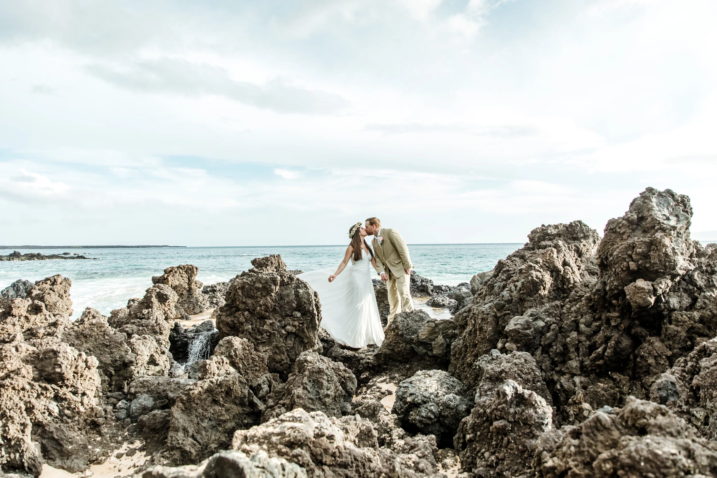 A bride and groom kissing on a rocky beach with the ocean in the background.
