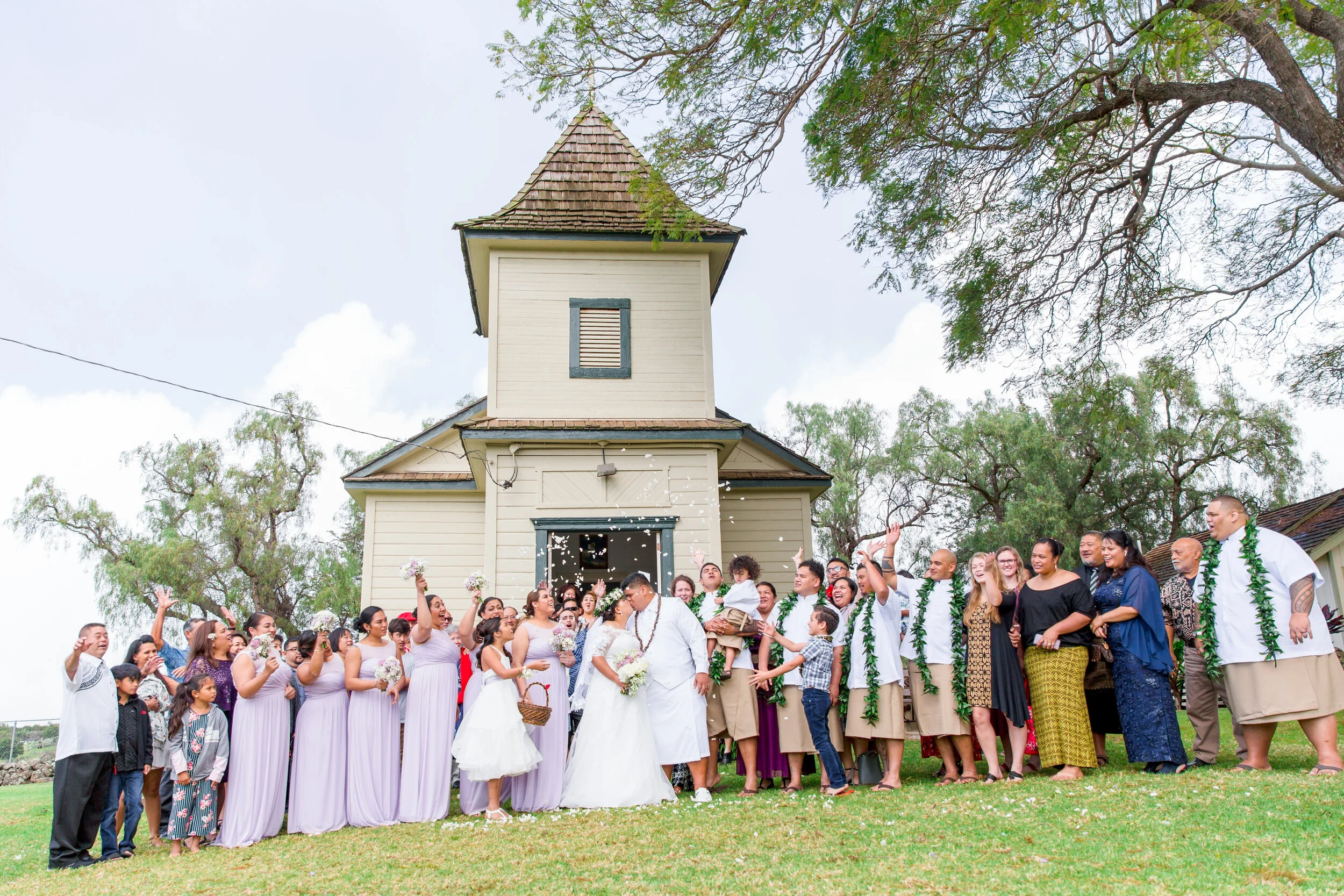 A large group of people dressed for a wedding celebration, gathered outside a church. The bride and groom are at the center, with the bride wearing a white dress and the groom in a white shirt. Guests are smiling, waving, and holding flowers, under a