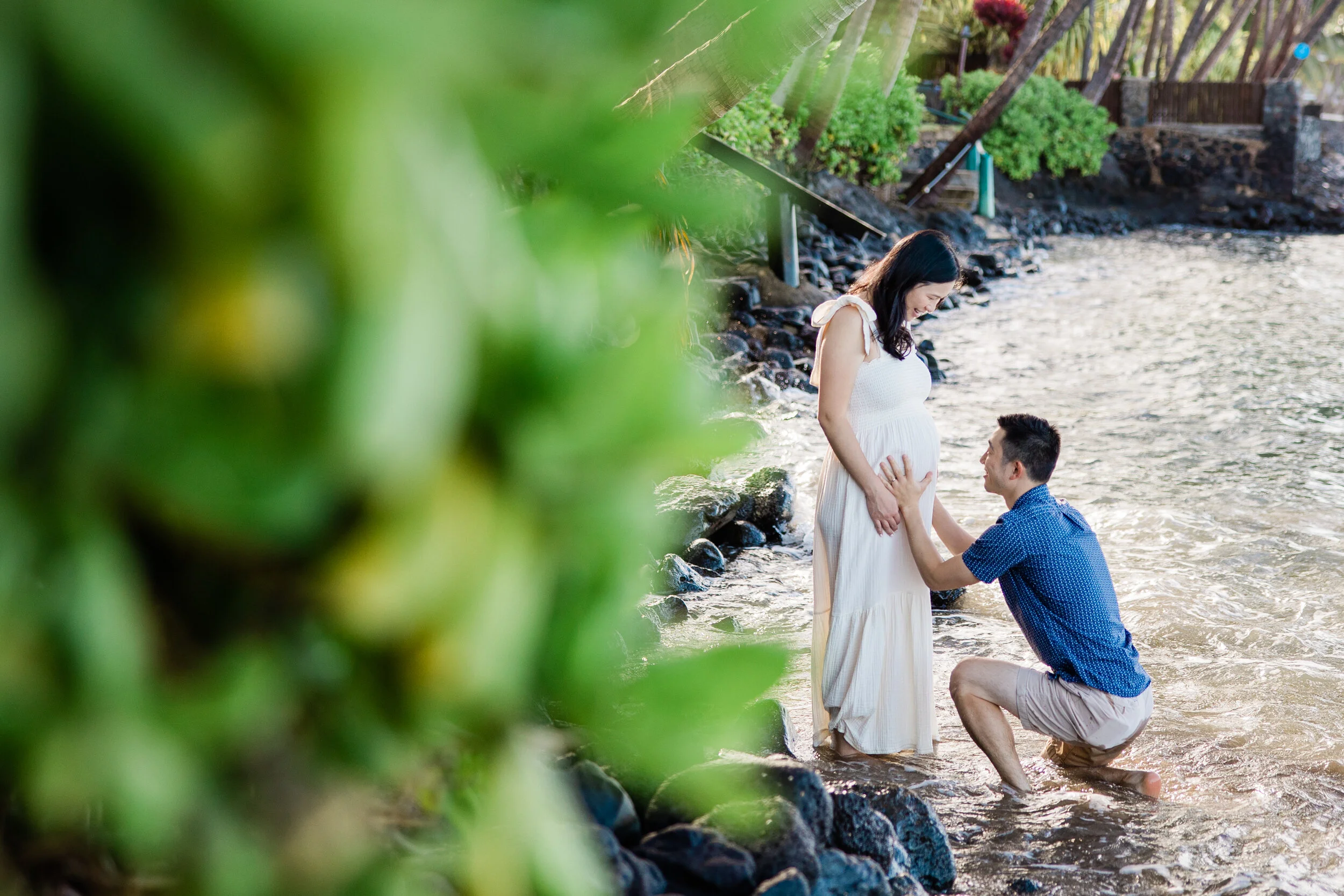 A pregnant woman in a white dress standing by the water's edge with a man kneeling before her, holding her hand, during sunset on a beach with greenery in the foreground.